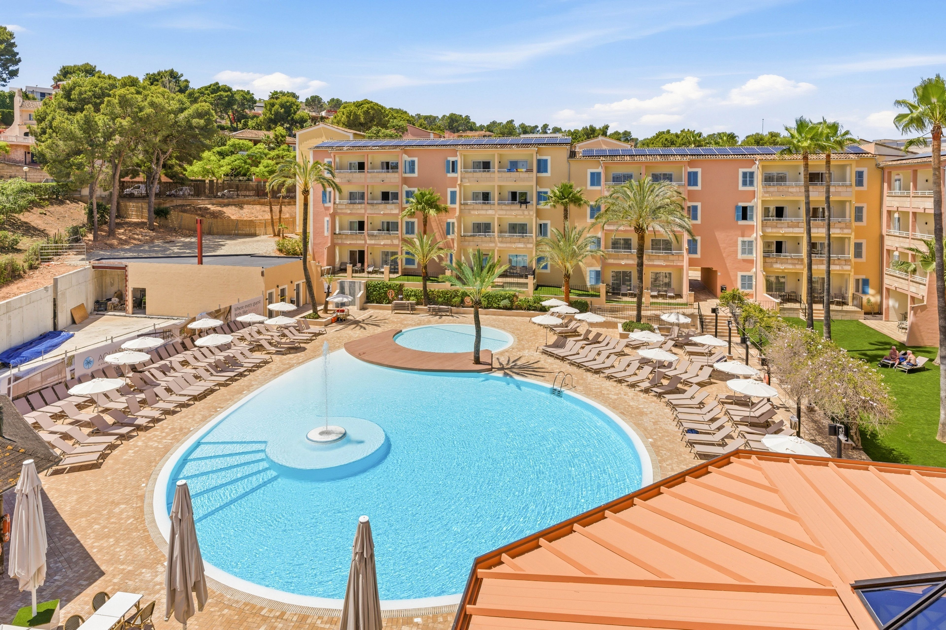 A large resort hotel features a curved outdoor swimming pool surrounded by rows of sun loungers and umbrellas. The multi-story building in the background has colorful facades with balconies overlooking the pool area. Palm trees and landscaped greenery add a tropical feel to the sunny courtyard. Clear blue skies and bright light suggest a warm vacation destination.