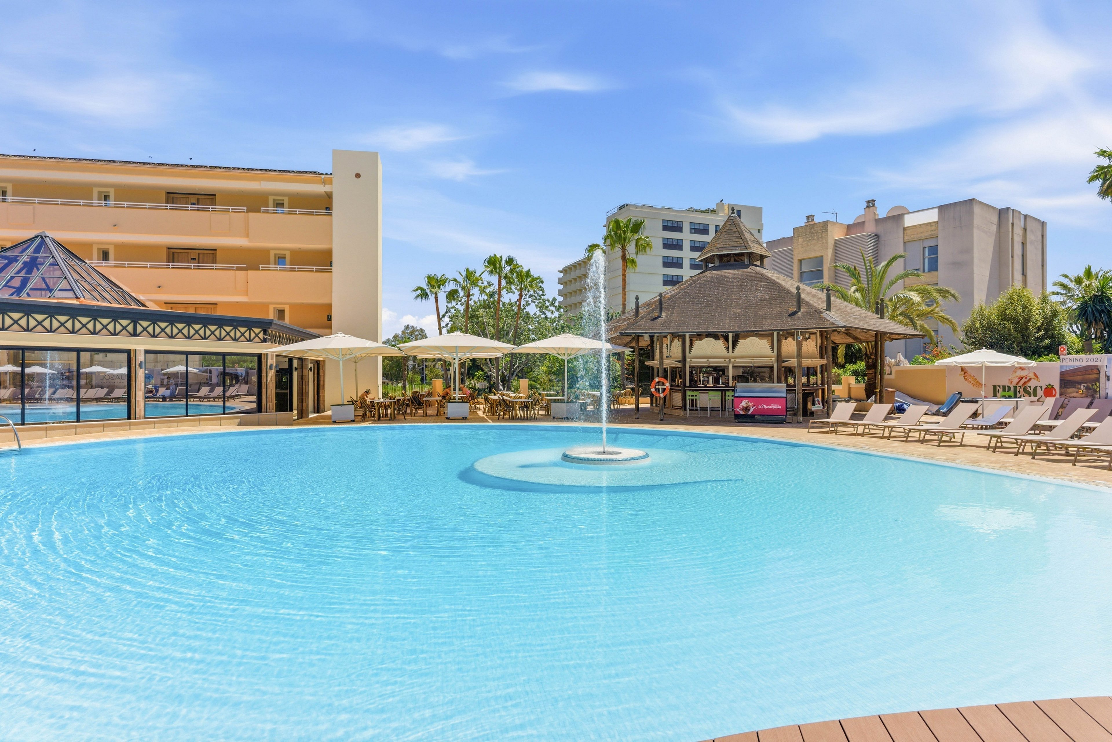 A large, clear blue swimming pool dominates the foreground of a sunny resort scene. Sun loungers and white umbrellas line the poolside, offering shaded relaxation areas. A thatched-roof pool bar and surrounding hotel buildings frame the background, with palm trees adding a tropical touch. The bright daylight and blue sky create a warm, inviting vacation atmosphere.