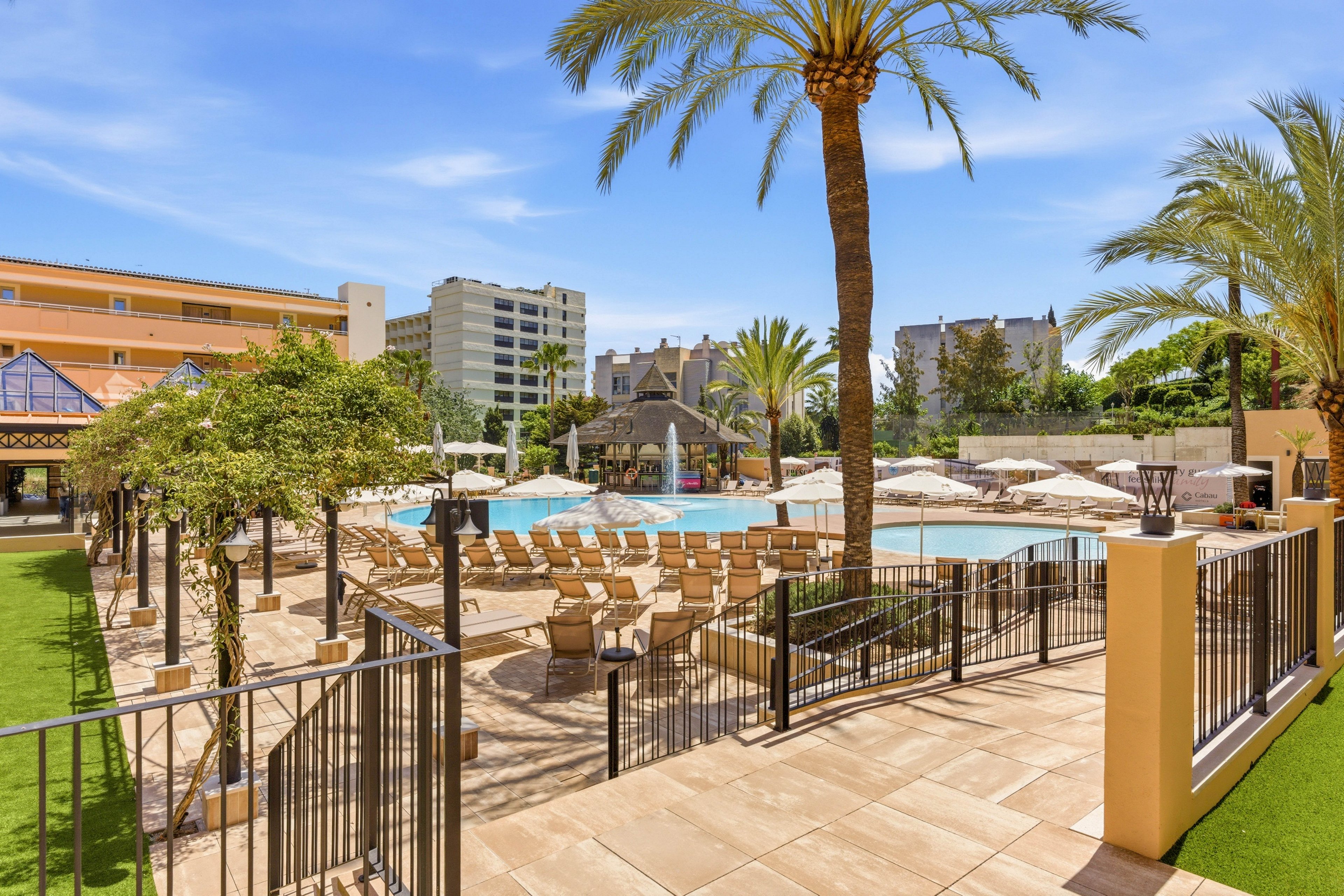 An outdoor resort swimming pool is surrounded by rows of empty sun loungers and umbrellas on a bright sunny day. Tall palm trees and manicured greenery frame the pool area, creating a tropical vacation atmosphere. Modern multi-story buildings and hotel structures rise in the background under a clear blue sky. Metal railings and tiled walkways lead toward the pool, emphasizing the organized and relaxing leisure setting.