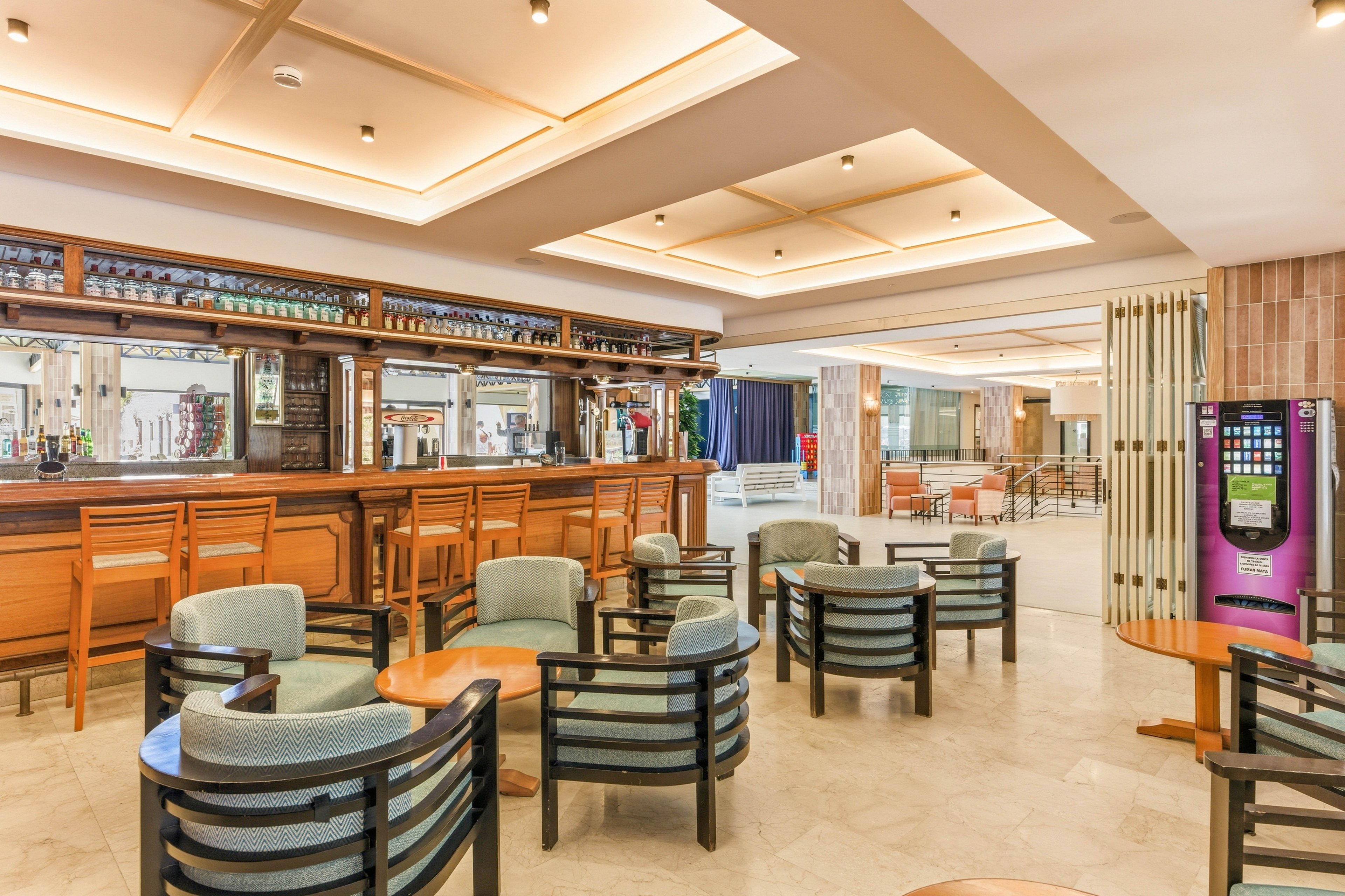 A spacious hotel lobby bar features a long wooden counter with high stools and a mirrored back bar. Several round tables with cushioned armchairs are arranged on a polished floor, creating an inviting seating area. The ceiling has recessed lighting in square panels, giving the space a warm, ambient glow. A colorful vending machine stands to the side, adding a modern touch to the otherwise classic interior.