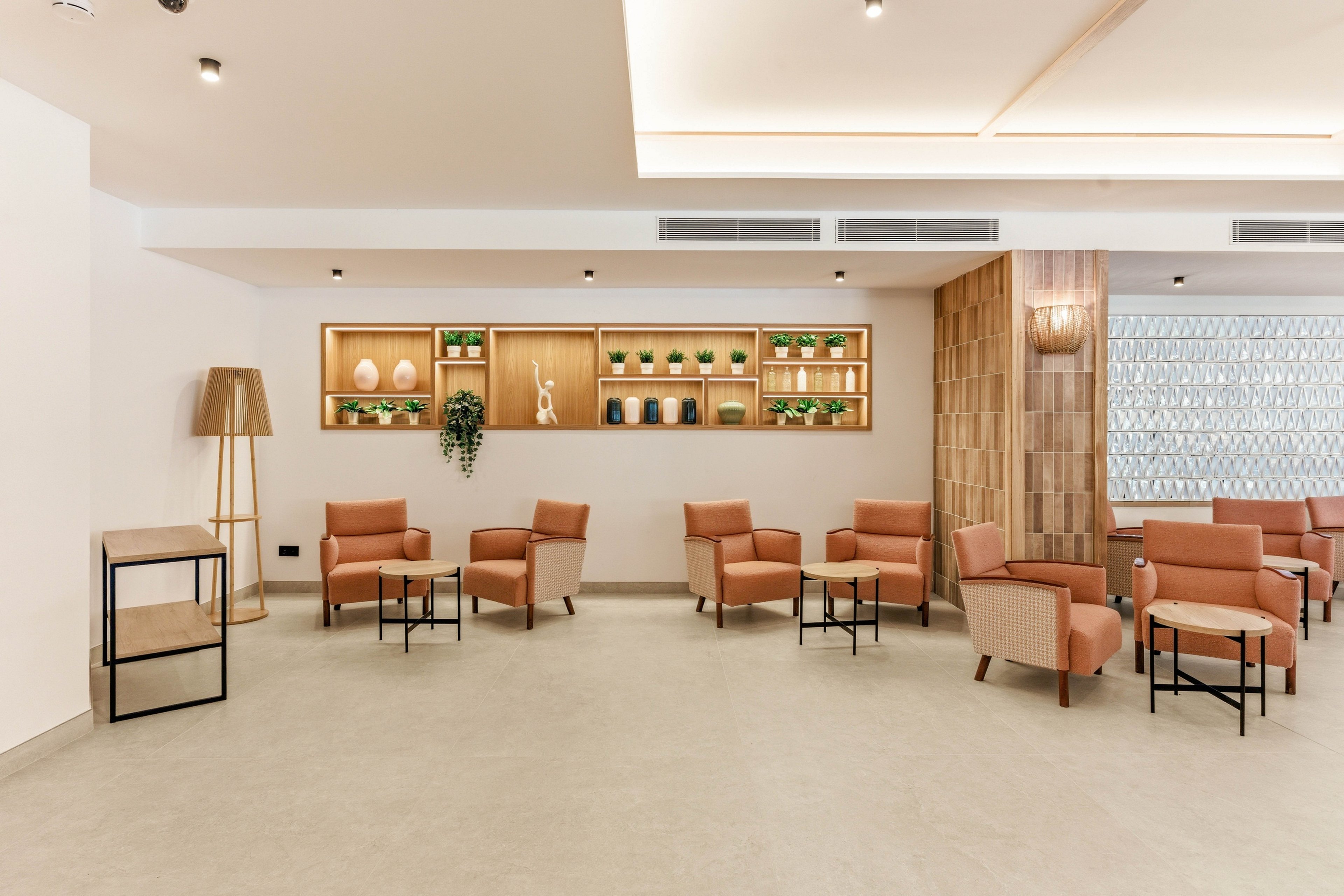 Contemporary lounge area featuring multiple tan armchairs arranged around small black metal tables. The setting is a bright, spacious interior with light walls and a neutral floor. A wooden shelving unit on the back wall displays plants, vases, and decorative objects. Warm lighting from ceiling fixtures and a floor lamp enhances the cozy, minimalist atmosphere.