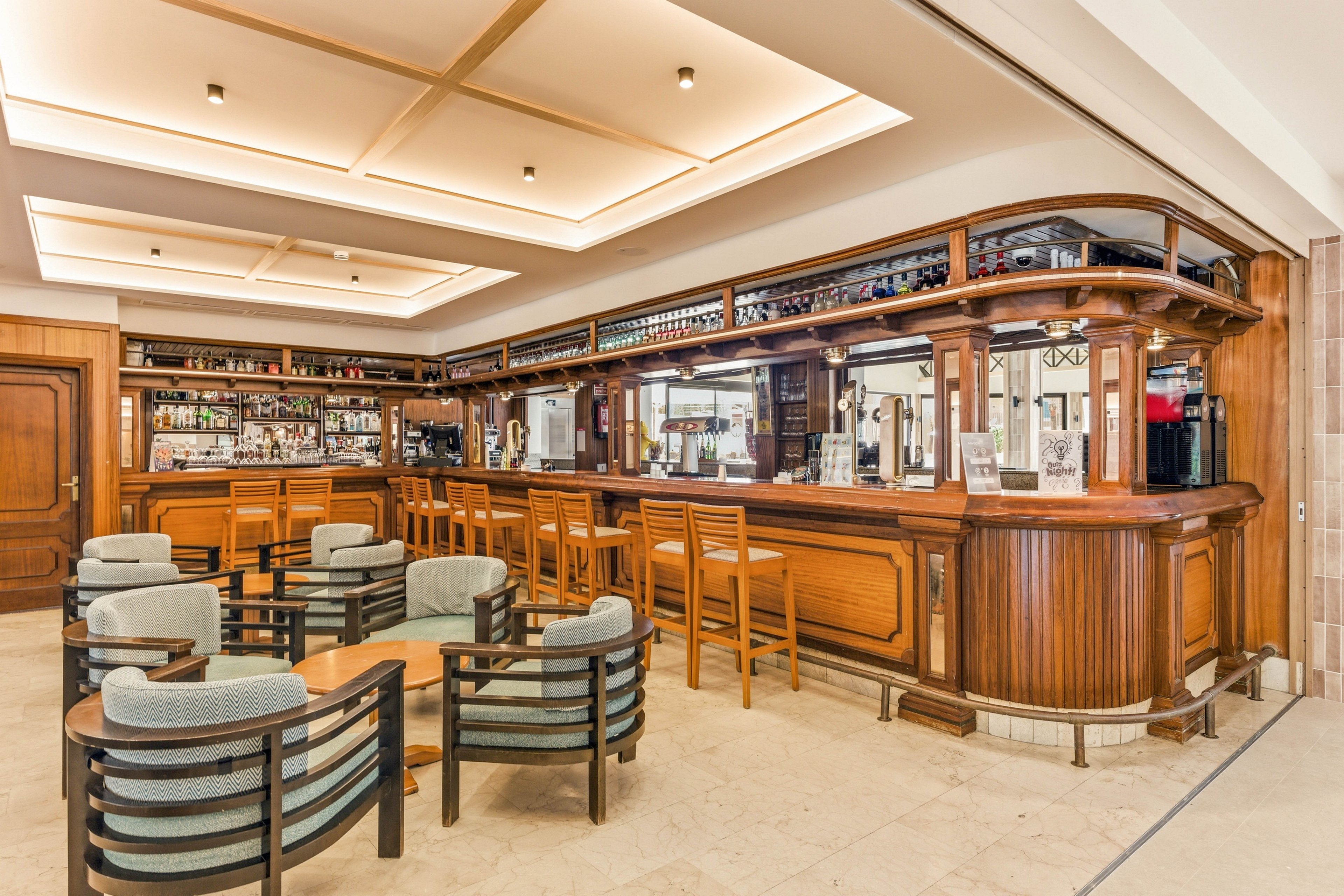 Spacious hotel bar interior featuring a long wooden counter and back bar shelving stocked with bottles and glassware. Several round tables with cushioned armchairs are arranged in the foreground, creating an inviting lounge area. The room is brightly lit with recessed ceiling panels and light-colored flooring. No people are present, emphasizing the clean, orderly, and tranquil atmosphere.