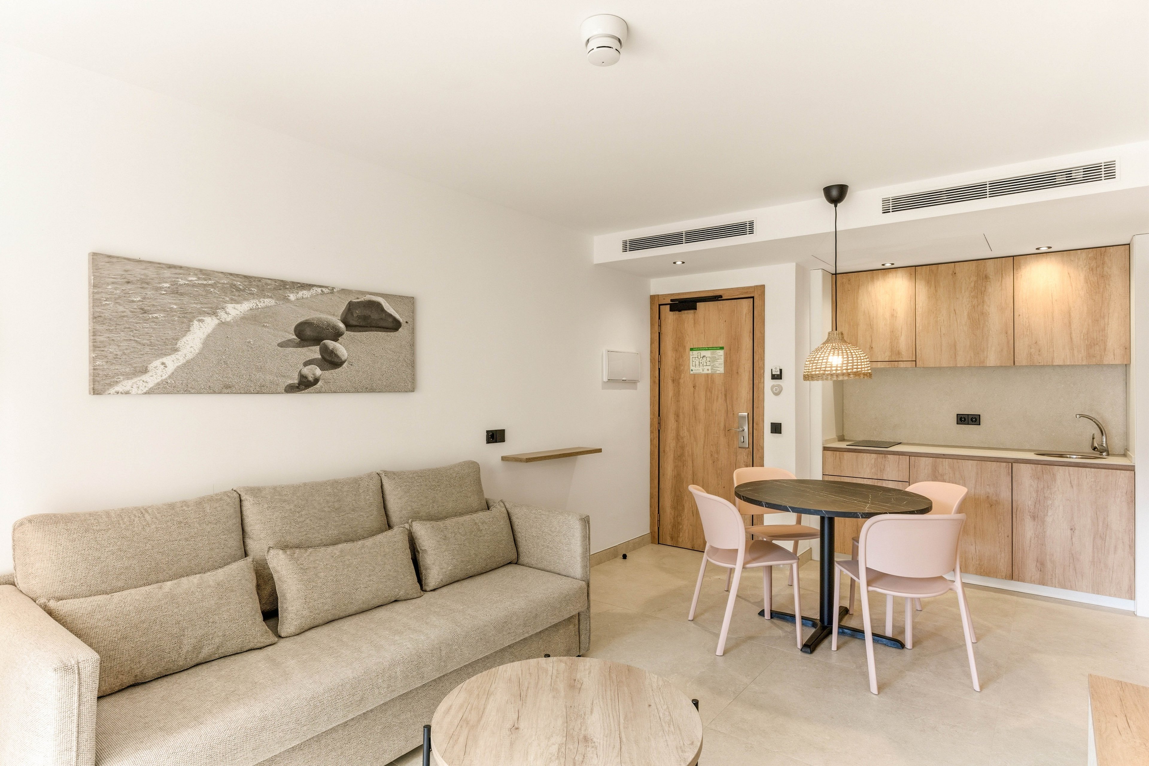 A bright modern studio apartment features a beige sofa and minimalist decor. The space includes a compact kitchenette with light wood cabinets, sink, and under-cabinet lighting. A small round dining table with pastel chairs sits near the entrance door. Neutral tones, clean lines, and simple artwork create a calm, contemporary atmosphere.