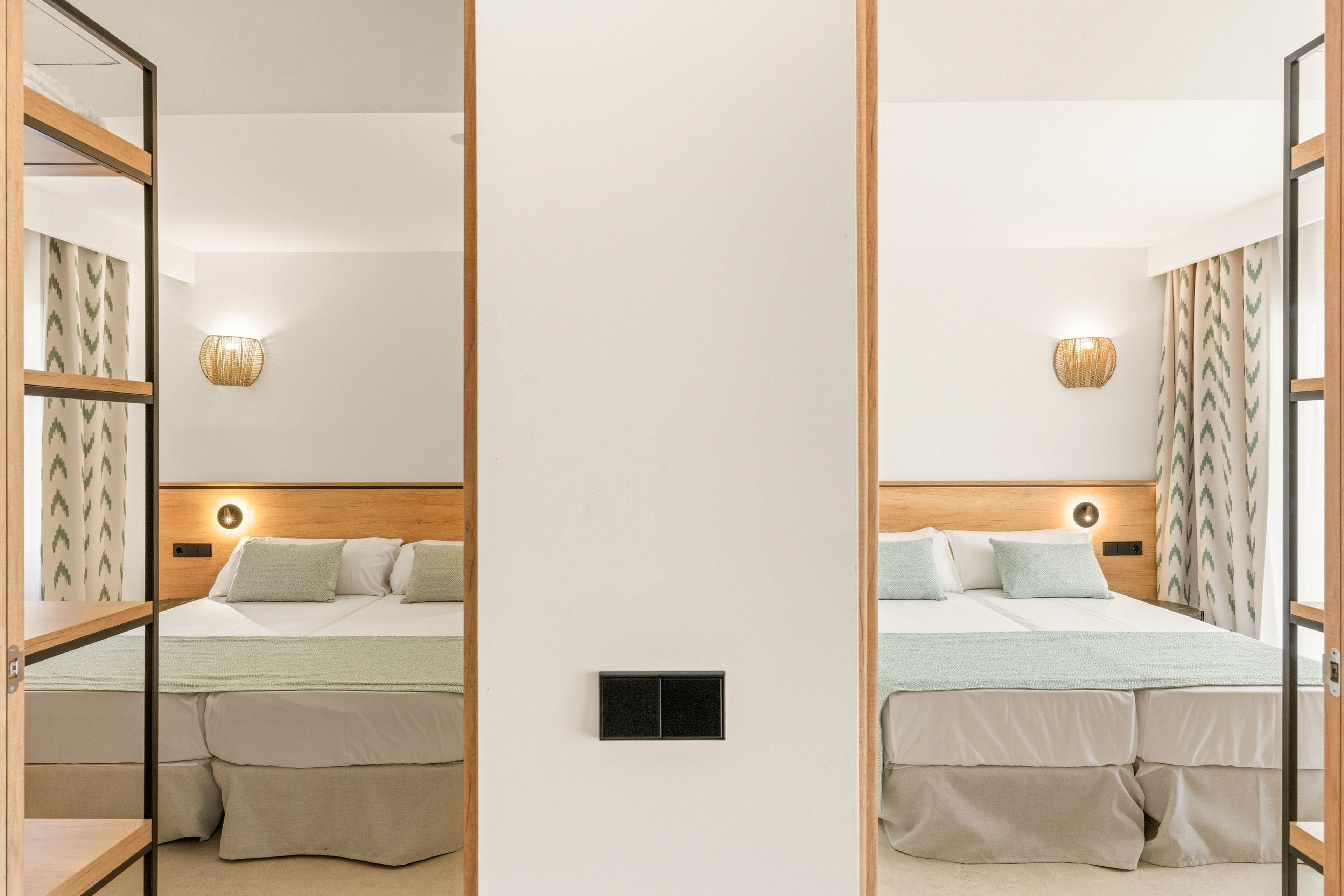 A contemporary hotel bedroom features a symmetrical layout with two identical beds on either side of a central wall. Soft neutral tones and light wood accents create a calm and inviting atmosphere. Wall-mounted lamps and patterned curtains add subtle decorative interest. The overall design emphasizes clean lines, balance, and minimalism.