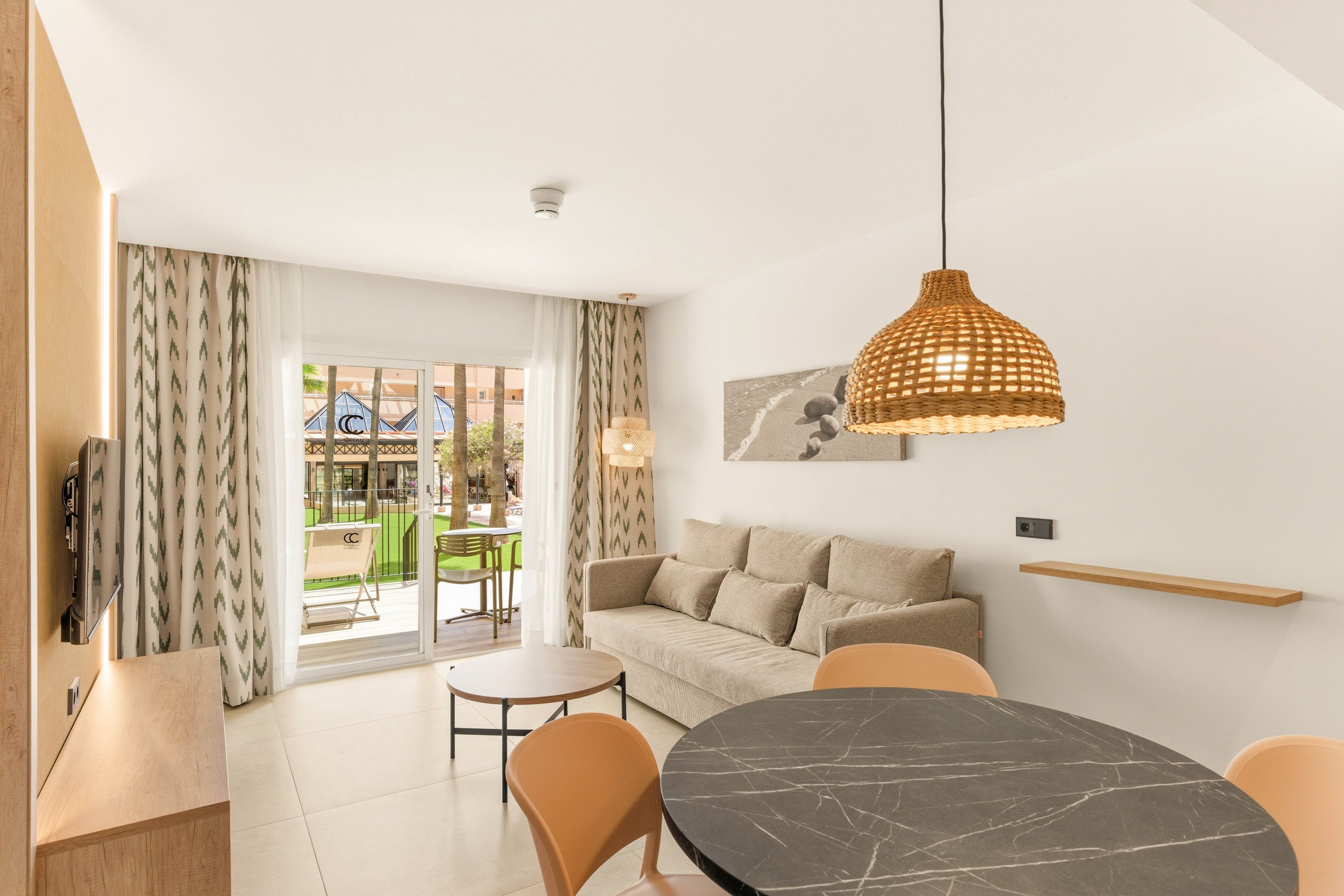 A contemporary living room features a beige sofa and a round black marble-style dining table with tan chairs. A woven rattan pendant light hangs from the ceiling, adding warmth to the neutral interior. Large glass doors open to a sunny balcony with green chairs and outdoor views. Light curtains, minimal wall art, and clean lines create an airy, modern atmosphere.