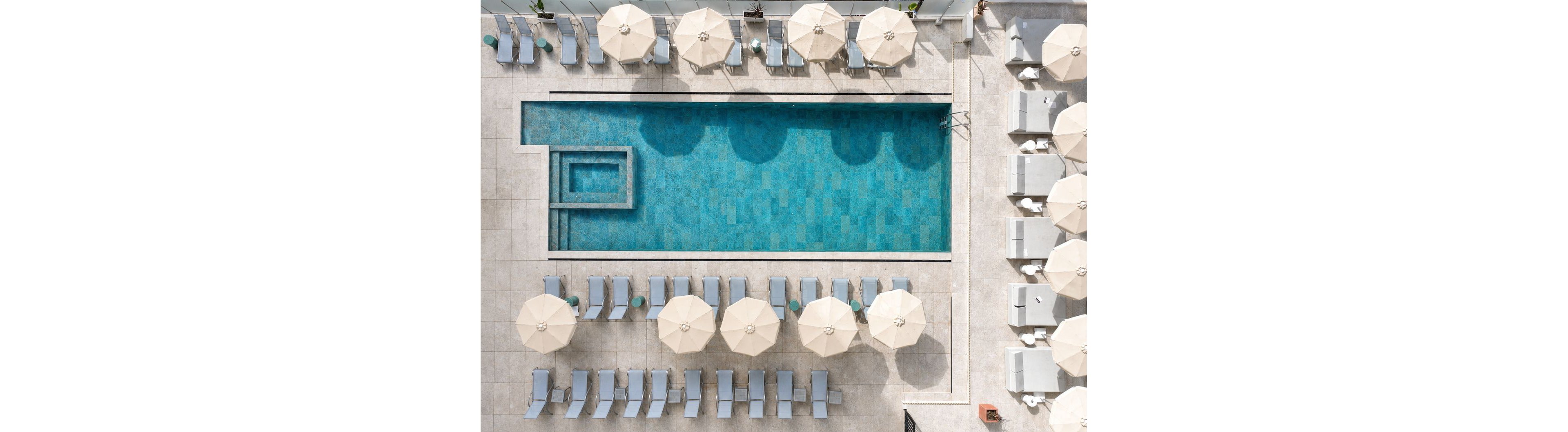 An aerial view shows a rectangular swimming pool surrounded by neatly arranged lounge chairs and umbrellas. The pool features a smaller square section, possibly a spa or shallow area, connected to the main pool. Light-colored tiles and blue water create a clean, modern aesthetic. The scene appears calm and organized, suggesting a resort or hotel rooftop setting.