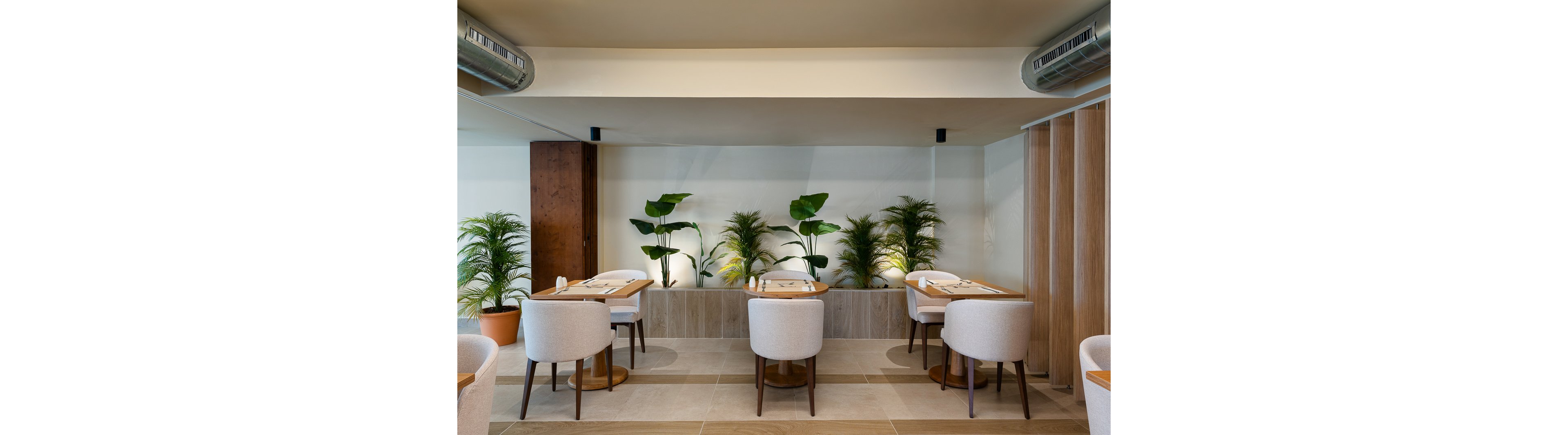 The image shows a contemporary restaurant dining area featuring light-colored upholstered chairs and wooden tables. The setting is enhanced by lush green plants arranged along the wall, creating a fresh and inviting atmosphere. The palette is neutral, with beige and natural wood accents, and the space is well-lit with soft lighting. No visible text or numeric values are present.