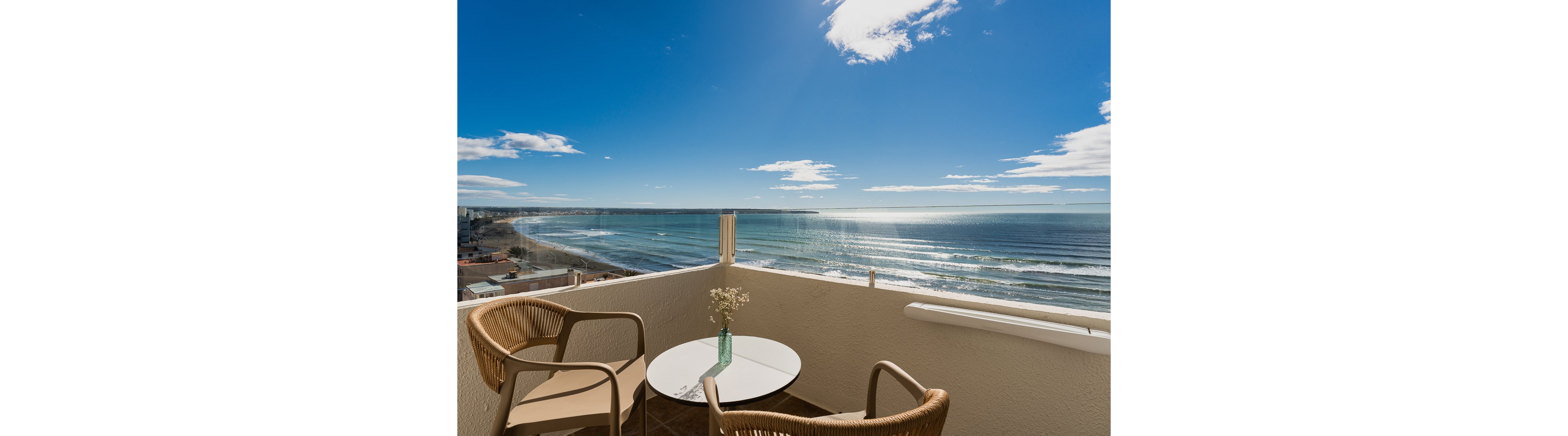 A bright balcony overlooks a wide expanse of ocean under a clear blue sky. Two wicker chairs and a small round table with a vase of flowers create a cozy seating area. Sunlight reflects off the water, and the coastline stretches into the distance, evoking a tranquil and inviting atmosphere.