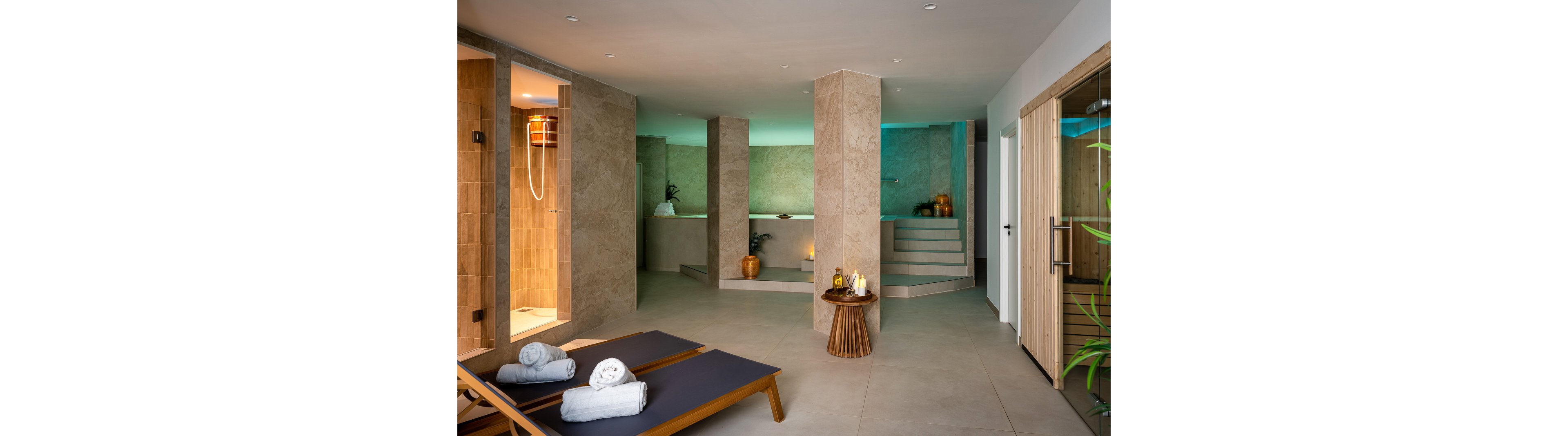 The image showcases a contemporary spa setting featuring two lounge chairs with rolled towels, a sauna room, and a tranquil ambiance. Neutral stone walls and soft lighting create a relaxing mood, while decorative elements like candles and plants enhance the atmosphere. Steps lead to a raised area, and the space is designed for wellness and comfort.
