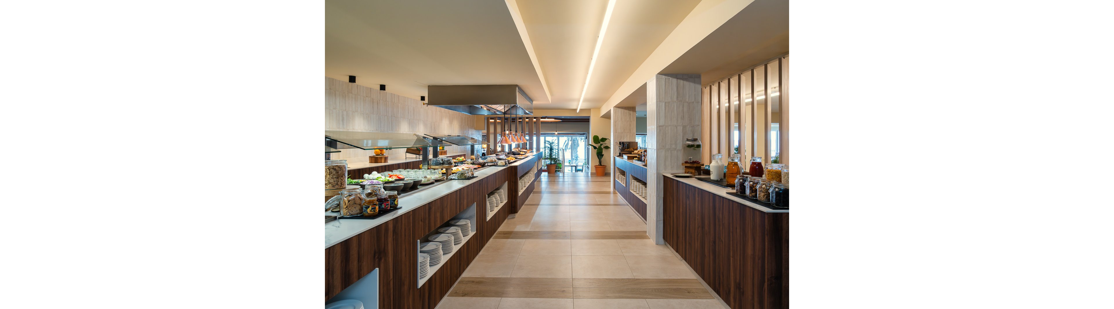 The image shows a spacious buffet restaurant featuring multiple food stations with a variety of dishes and beverages. The setting is contemporary, with wood and tile finishes, open counters, and neatly arranged plates and jars. Natural light streams in from large windows at the far end, creating a welcoming and bright atmosphere. No visible text or numeric values are present in the image.