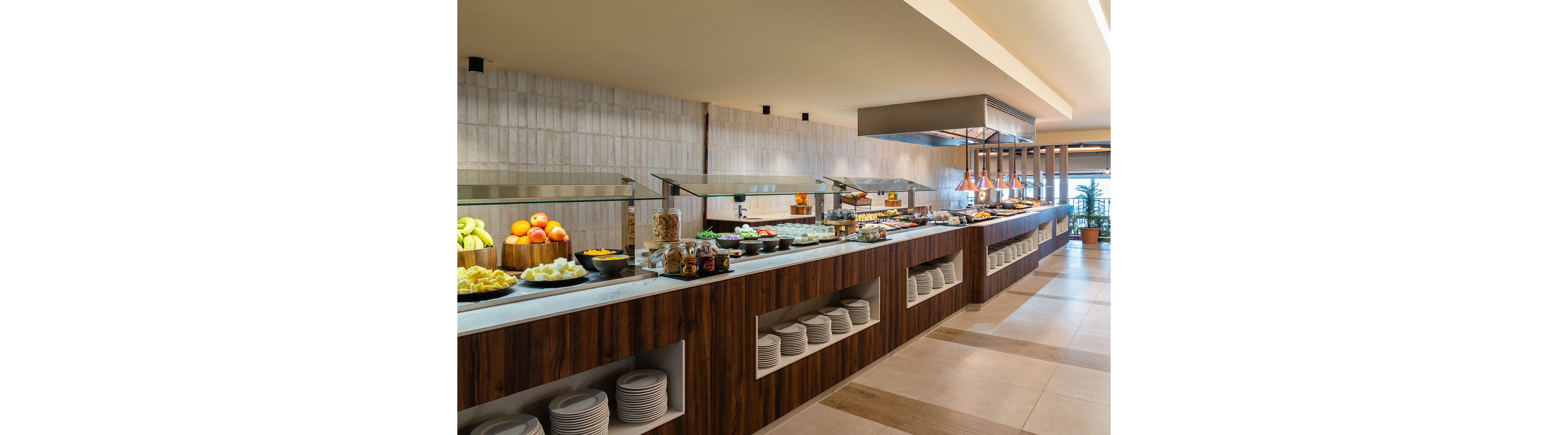 A spacious buffet counter displays a variety of breakfast items, including fresh fruits, cereals, and pastries. The setting is a contemporary dining area with warm lighting and wooden accents, creating a welcoming atmosphere. Plates and bowls are neatly stacked below the counter, and the food is arranged in an organized manner. The overall mood is inviting and clean, ideal for hotel guests seeking a morning meal.