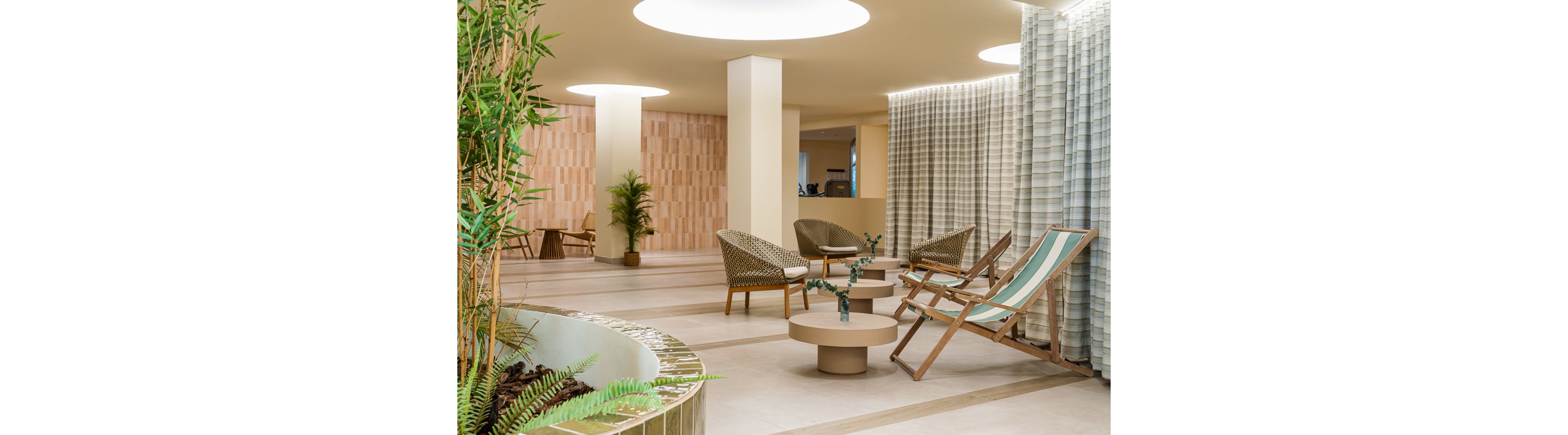 A contemporary lounge space features wicker chairs, a striped deck chair, and round coffee tables. The setting is indoors with soft beige tones, natural wood accents, and lush green plants adding a touch of freshness. Large circular ceiling lights and sheer curtains contribute to a calm, inviting atmosphere. The area is empty, emphasizing relaxation and modern design.