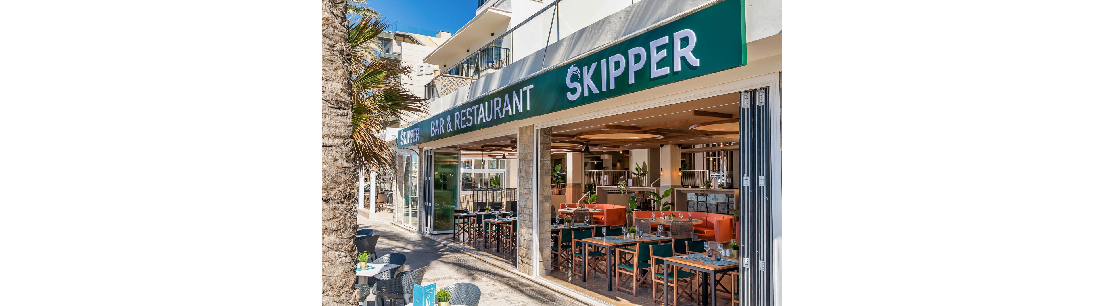 The image shows the exterior of Skipper Bar & Restaurant, featuring a modern open-air dining space with tables and chairs set for guests. The setting is bright and sunny, with palm trees and contemporary architecture visible. The restaurant's name 'SKIPPER' and 'BAR & RESTAURANT' are clearly displayed on the green signage above the entrance. The mood is inviting and relaxed, ideal for casual dining.