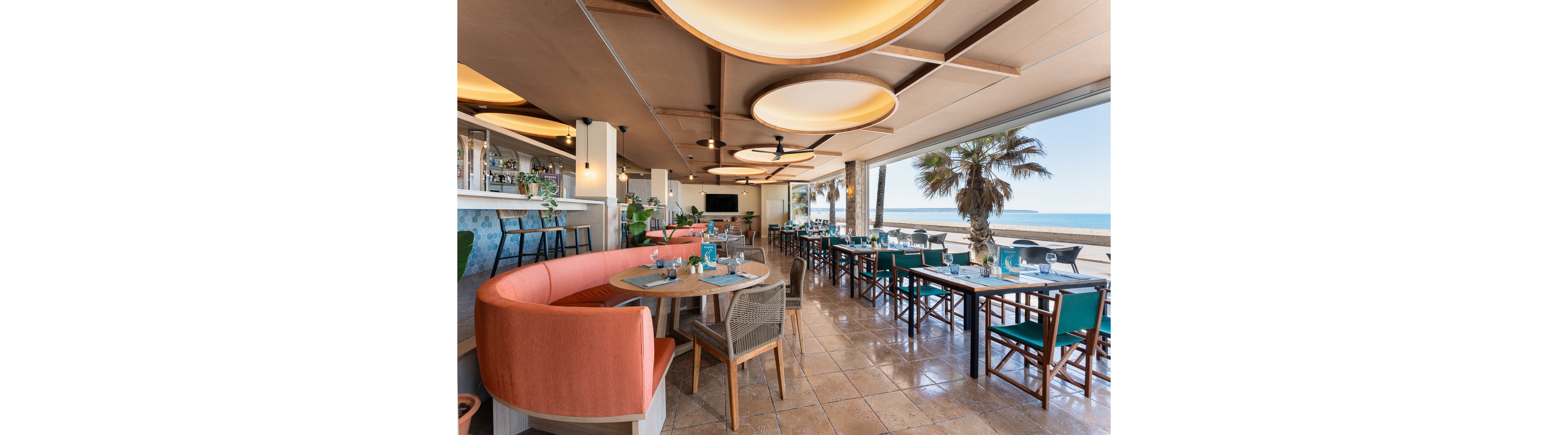 The image shows a contemporary restaurant interior featuring stylish seating arrangements, including curved booths and wooden chairs. Large windows open to a scenic beach and ocean view, with palm trees visible outside. The setting is bright and airy, with turquoise accents and neatly set tables, creating a relaxed coastal atmosphere. No visible text or numeric values are present in the image.
