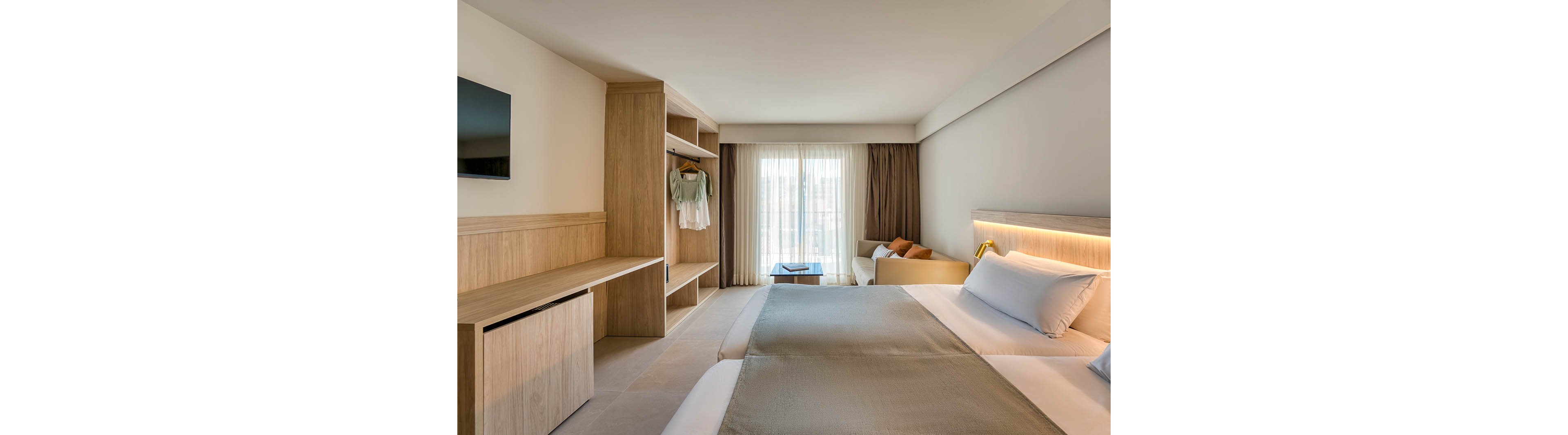 A contemporary hotel room features a double bed with neutral bedding, a wall-mounted television, and an open wardrobe with hanging clothes. Sunlight streams through sheer curtains covering a large window, illuminating the minimalist decor and light wood furnishings. A small sitting area with a sofa and coffee table is visible near the window, creating a welcoming and comfortable atmosphere.