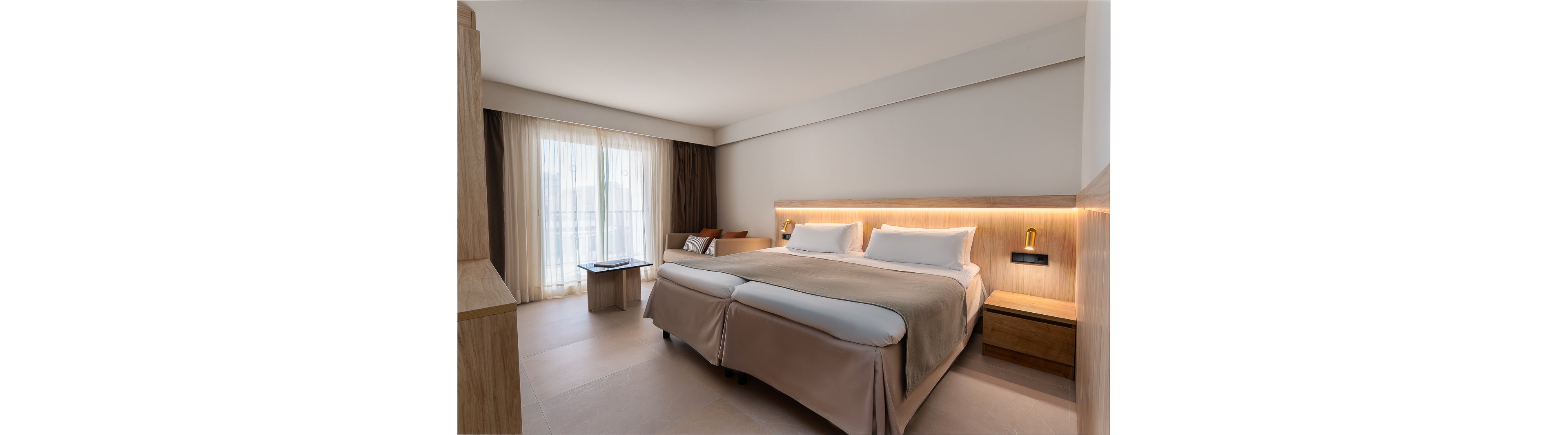 A contemporary hotel room features two twin beds with crisp white linens and beige covers. The space is illuminated by natural light streaming through large windows with sheer and blackout curtains. Minimalist wooden furniture, including a bedside table and a small seating area, complements the neutral palette. Warm ambient lighting above the headboard adds a cozy touch to the clean, modern design.