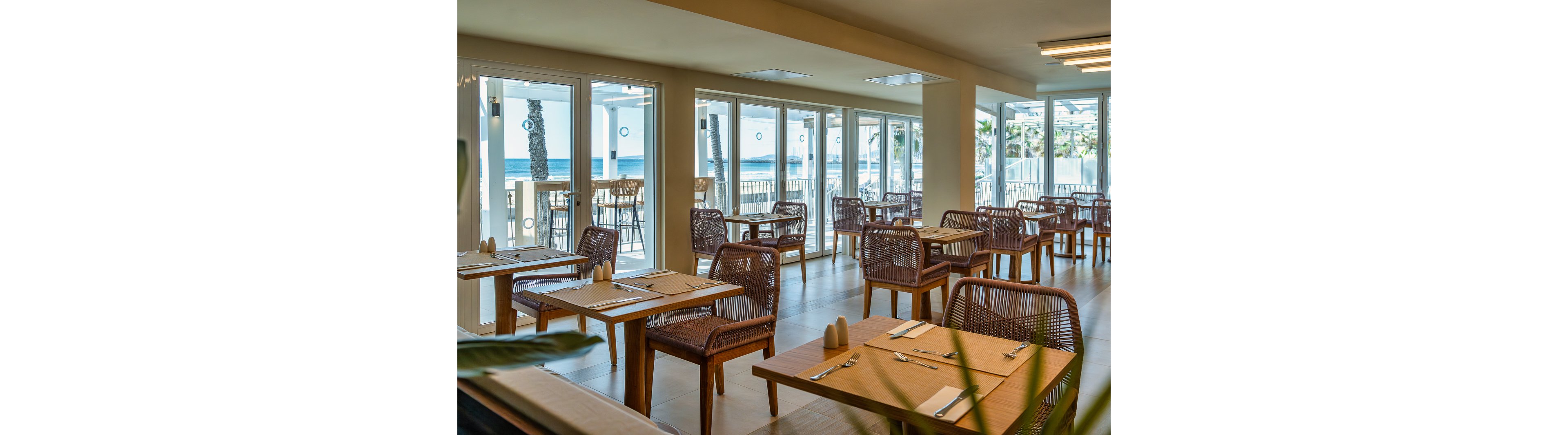 A modern restaurant dining area features wooden tables and wicker chairs, set with placemats and cutlery. Large windows provide a clear view of the ocean and palm trees outside, filling the space with natural light. The setting is calm and inviting, with a neutral color palette and a relaxed coastal atmosphere. No visible text or numbers are present in the image.