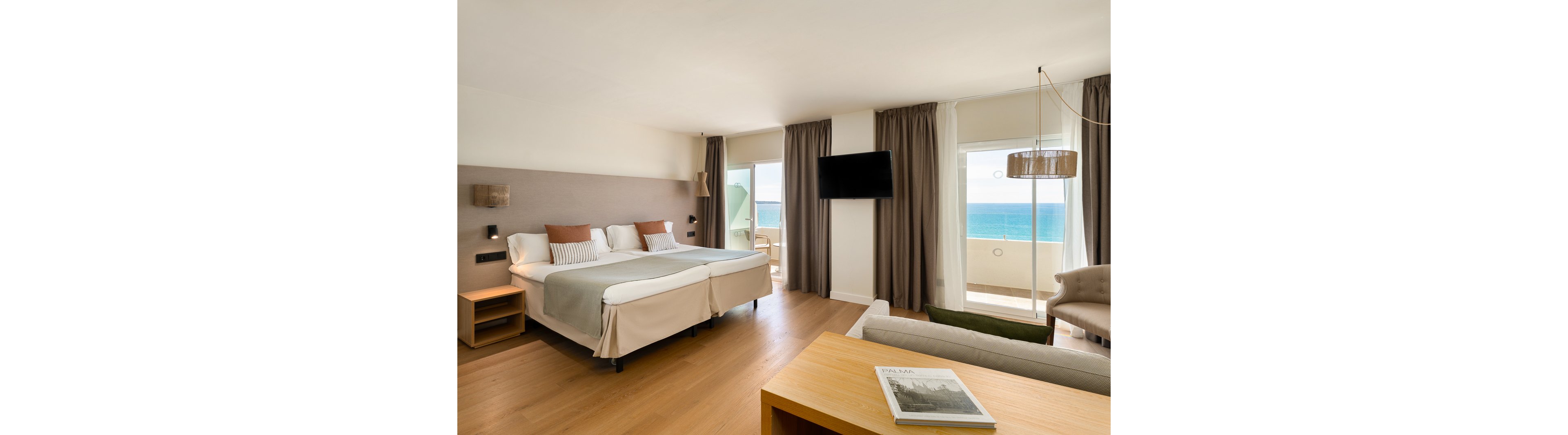 A spacious hotel room features two twin beds, a wall-mounted television, and a seating area with a sofa and armchair. Large windows and a glass door open to a balcony overlooking the ocean, filling the space with natural light. The room is decorated in neutral tones with brown and beige accents, creating a calm and inviting atmosphere. A magazine is placed on the wooden desk in the foreground.