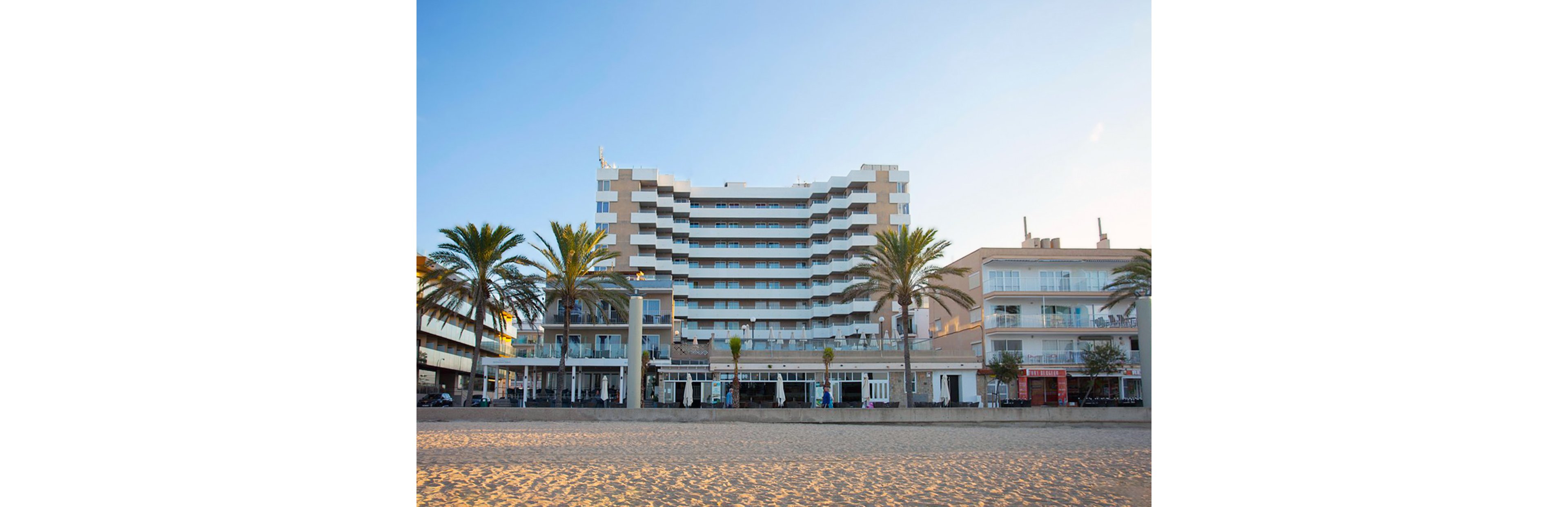A contemporary multi-story hotel stands directly on a sandy beach, framed by tall palm trees. The building features balconies and large windows, with a clear blue sky overhead and soft morning sunlight illuminating the scene. The foreground shows clean sand, and the hotel’s architecture is prominent, creating a relaxed coastal atmosphere.