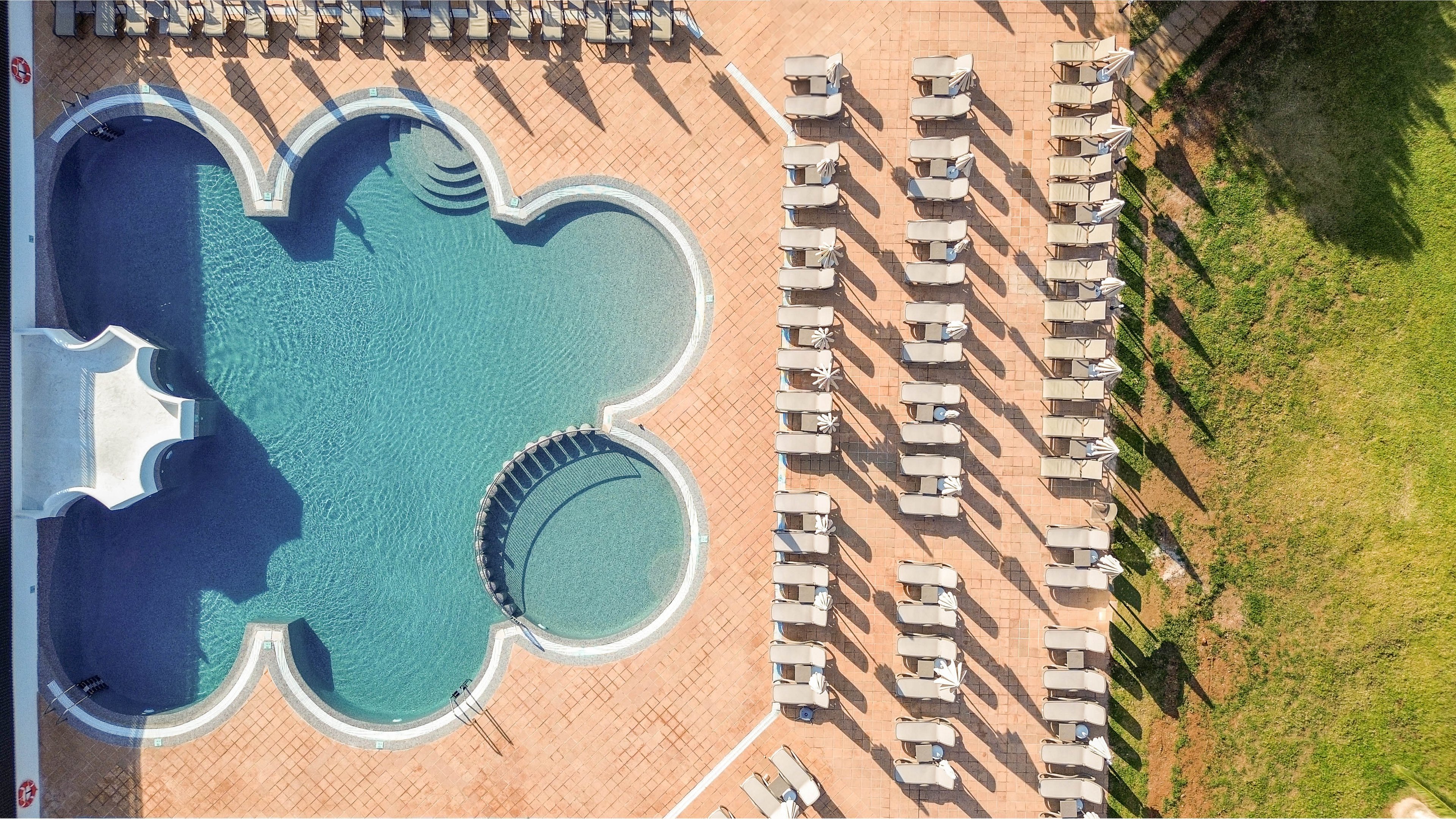 A uniquely shaped outdoor swimming pool is photographed from above at a resort. Rows of empty sun loungers line the terracotta-tiled pool deck, casting long shadows in the sunlight. A curved set of pool steps leads into the clear blue water, emphasizing the geometric design. A strip of green grass borders the deck, adding contrast to the warm and cool tones.