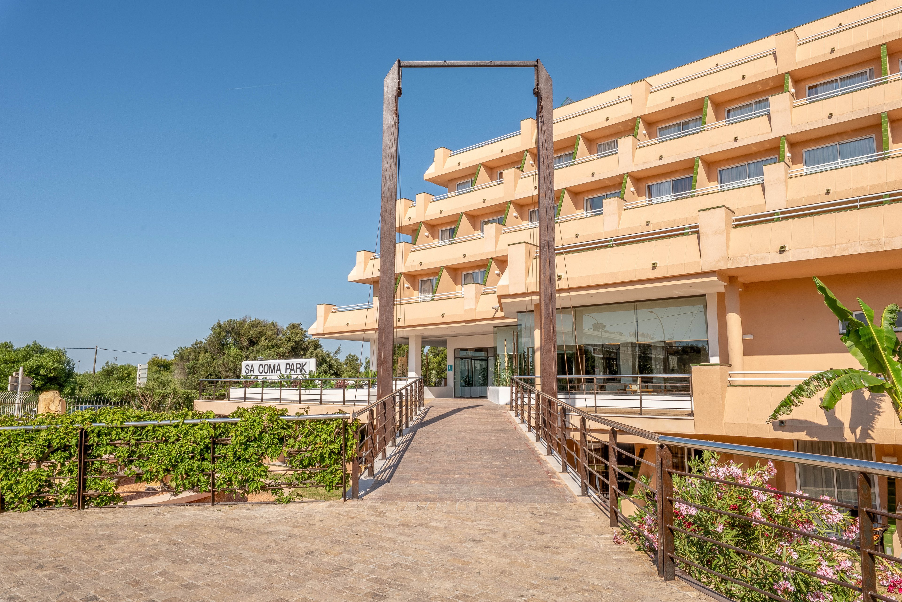 A modern multi-story resort hotel is shown under a clear blue sky. A wide pedestrian bridge with metal railings leads directly to the main glass entrance. The building features warm beige tones, balconies, and large windows. Green plants and shrubs line the sides of the bridge, adding a touch of nature to the contemporary architecture.