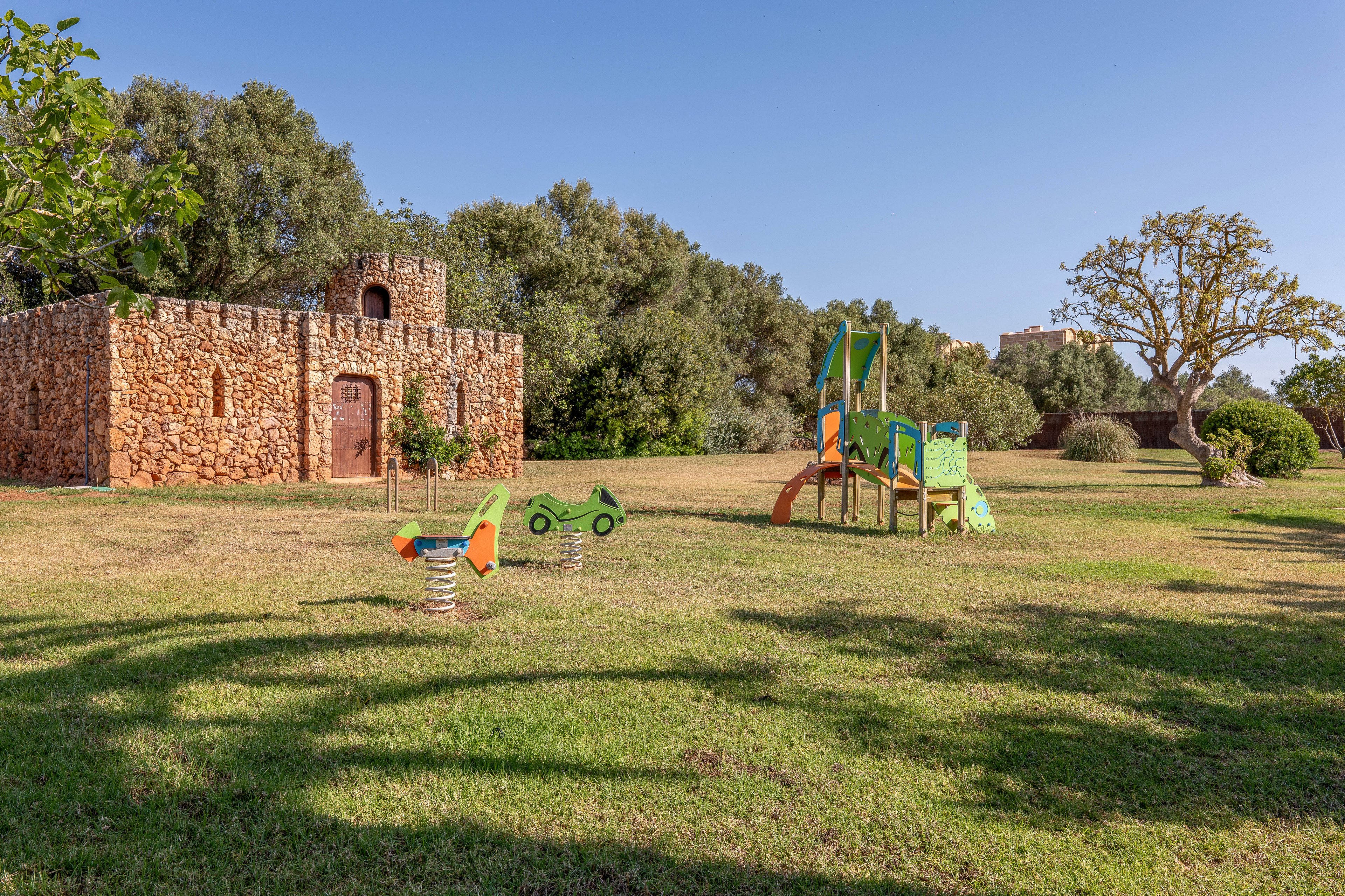 An outdoor playground stands on a wide grassy lawn under a clear blue sky. Colorful spring riders and a small slide structure are set in the open field. To the left, a rustic stone building with an arched wooden door and tower adds character to the scene. Mature trees and shrubs surround the area, creating a peaceful, family-friendly park setting.