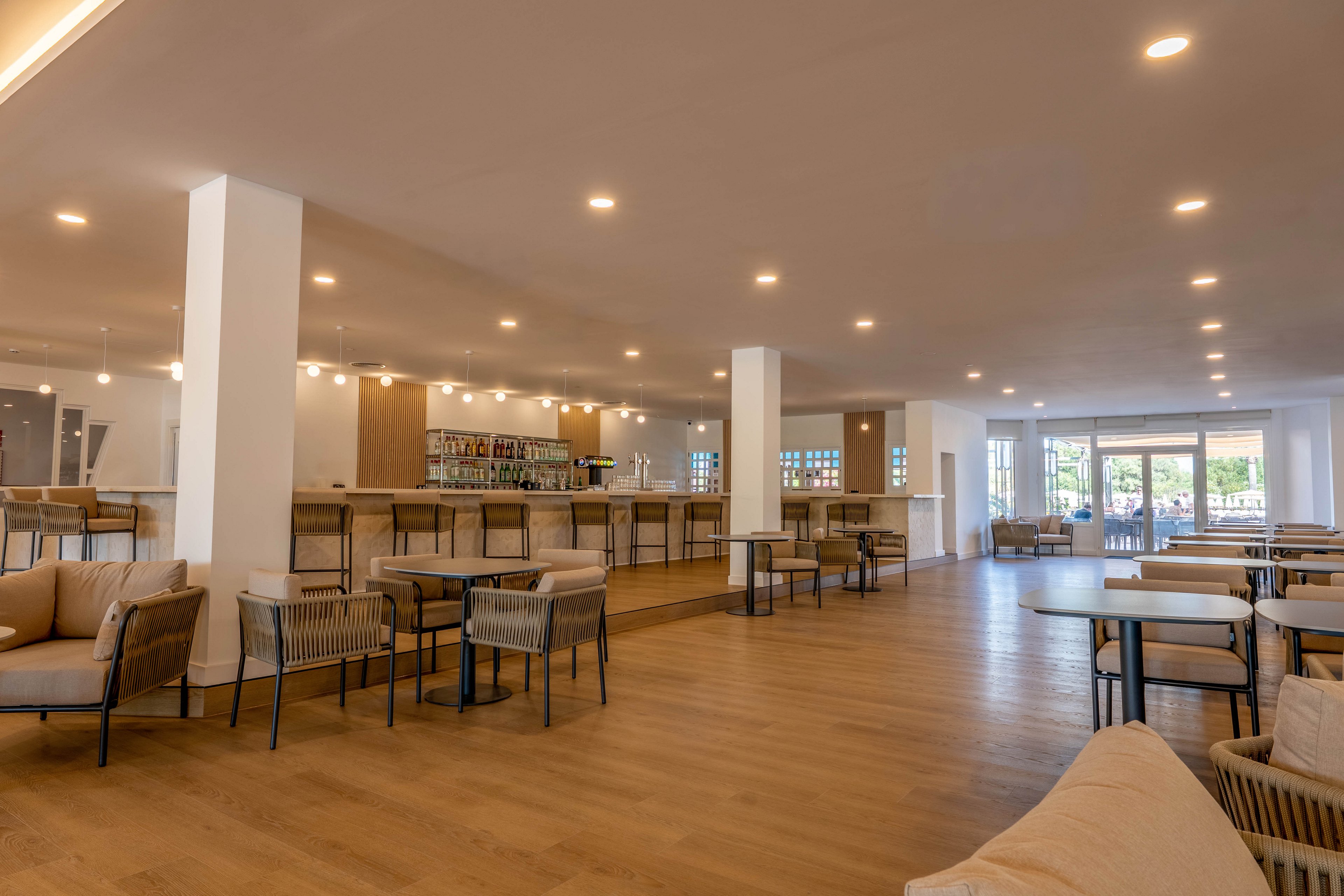 A spacious, contemporary bar lounge features a long counter with barstools and neatly arranged bottles in the background. Comfortable armchairs and tables are spread across a large wooden floor, creating an open seating area. Warm recessed ceiling lights and natural light from large windows brighten the room. The overall design is minimalist and inviting, suitable for socializing or relaxing in a hotel or resort setting.