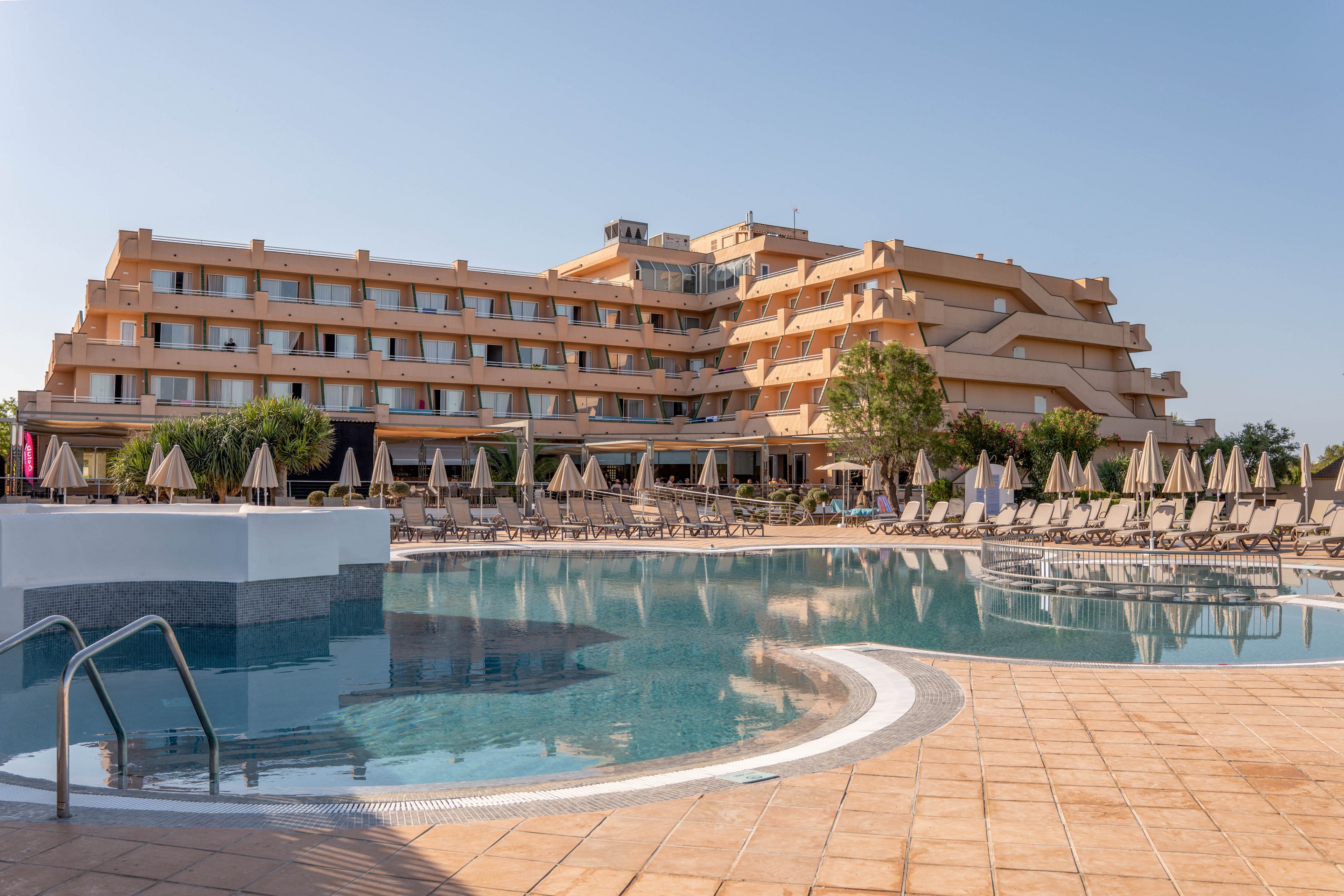 A large multi-story resort hotel overlooks a spacious outdoor swimming pool. Numerous sun loungers and umbrellas are arranged neatly around the poolside terrace. The scene is captured on a clear, sunny day with a bright blue sky. The overall atmosphere suggests a relaxing vacation destination focused on leisure and comfort.
