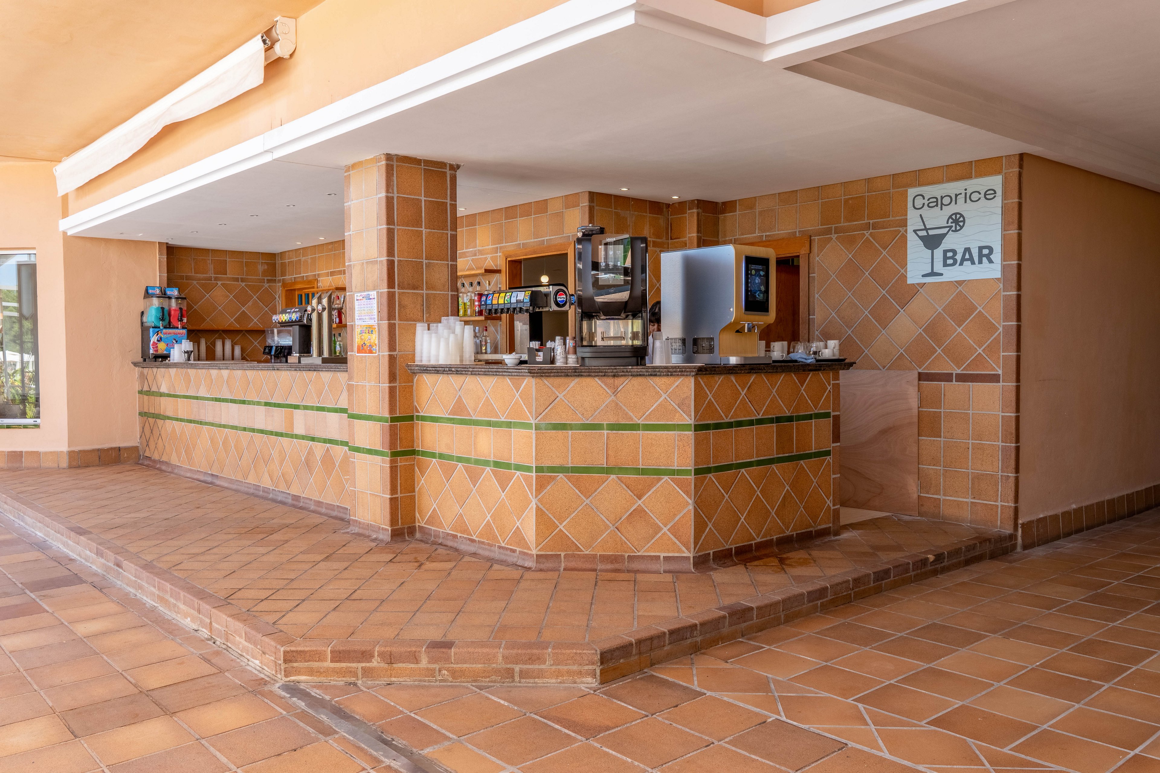An open-air bar counter with tiled surfaces stands ready for guests at a resort. Multiple beverage dispensers, coffee machines, and stacked plastic cups line the countertop. Warm terracotta tiles cover the floor, pillars, and bar front, creating a cohesive Mediterranean feel. A wall sign reading "Caprice BAR" with a martini glass icon identifies the bar area.
