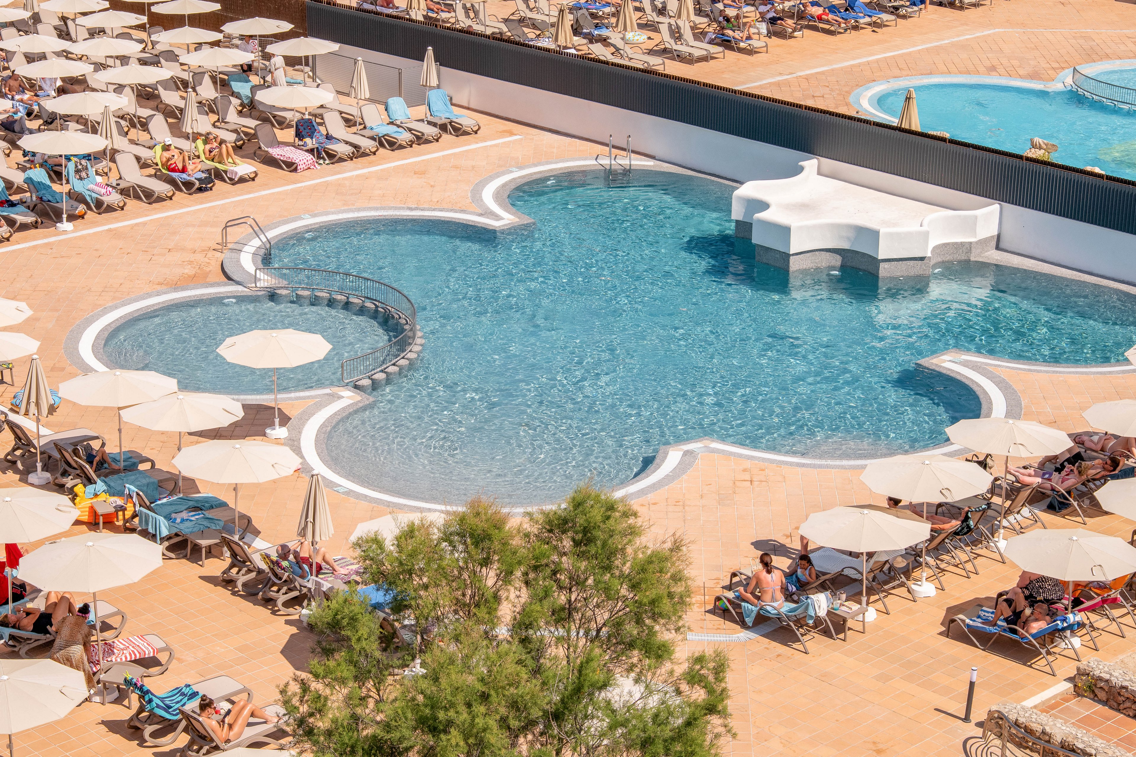 An aerial view shows a uniquely shaped resort swimming pool surrounded by numerous sun loungers and umbrellas. Guests relax on chairs and sunbathe around the pool deck in bright daylight. The pool features curved edges, a small bridge, and a white central platform structure. Warm tones of the tiled terrace contrast with the cool blue water, creating a summery vacation atmosphere.