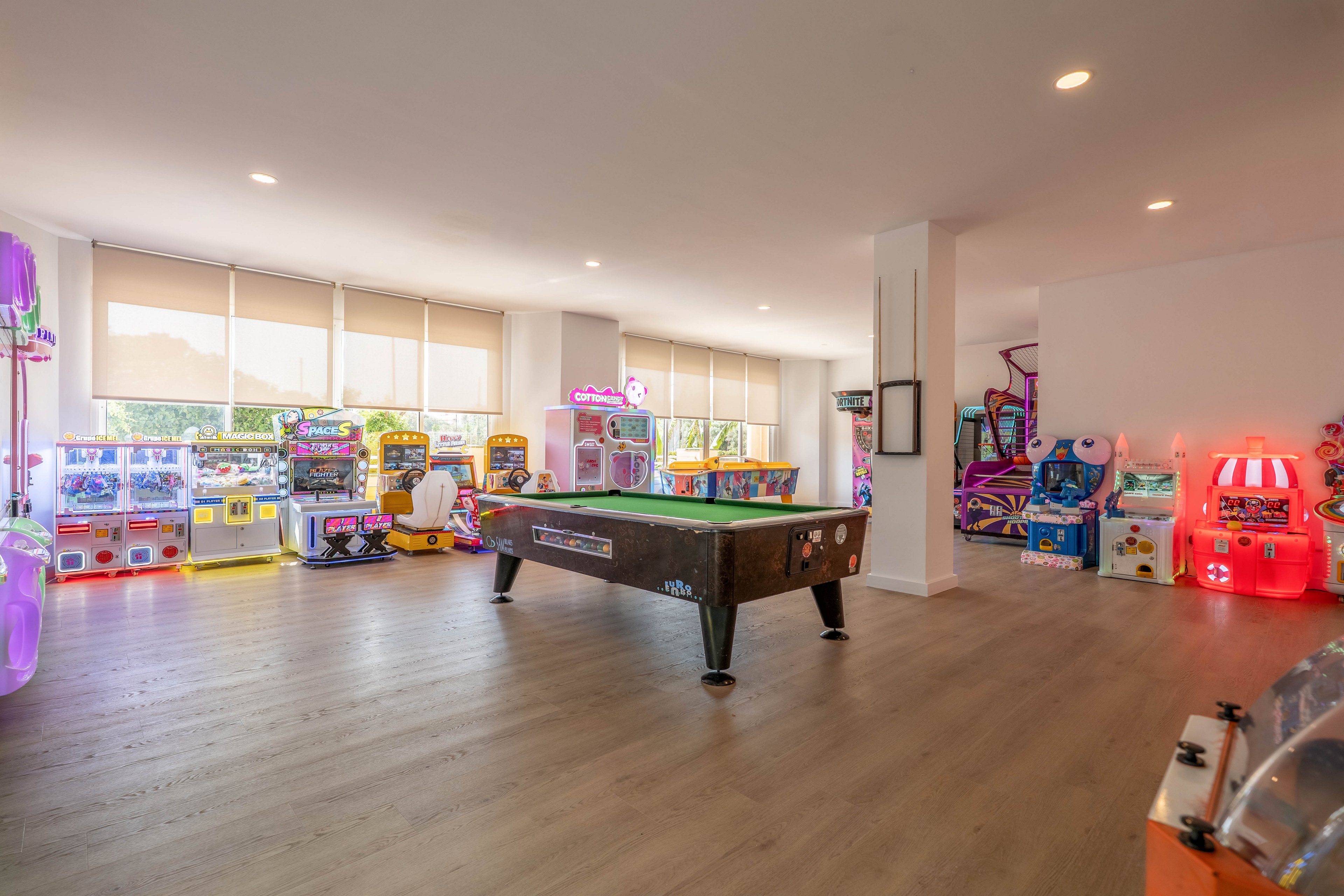 Spacious indoor game room featuring a pool table in the center. Colorful arcade machines and claw games line the walls, creating a playful entertainment area. Large windows with roller blinds let in natural light, softening the glow of neon lights from the machines. The clean, open layout and light wood flooring give the space a modern, inviting feel.