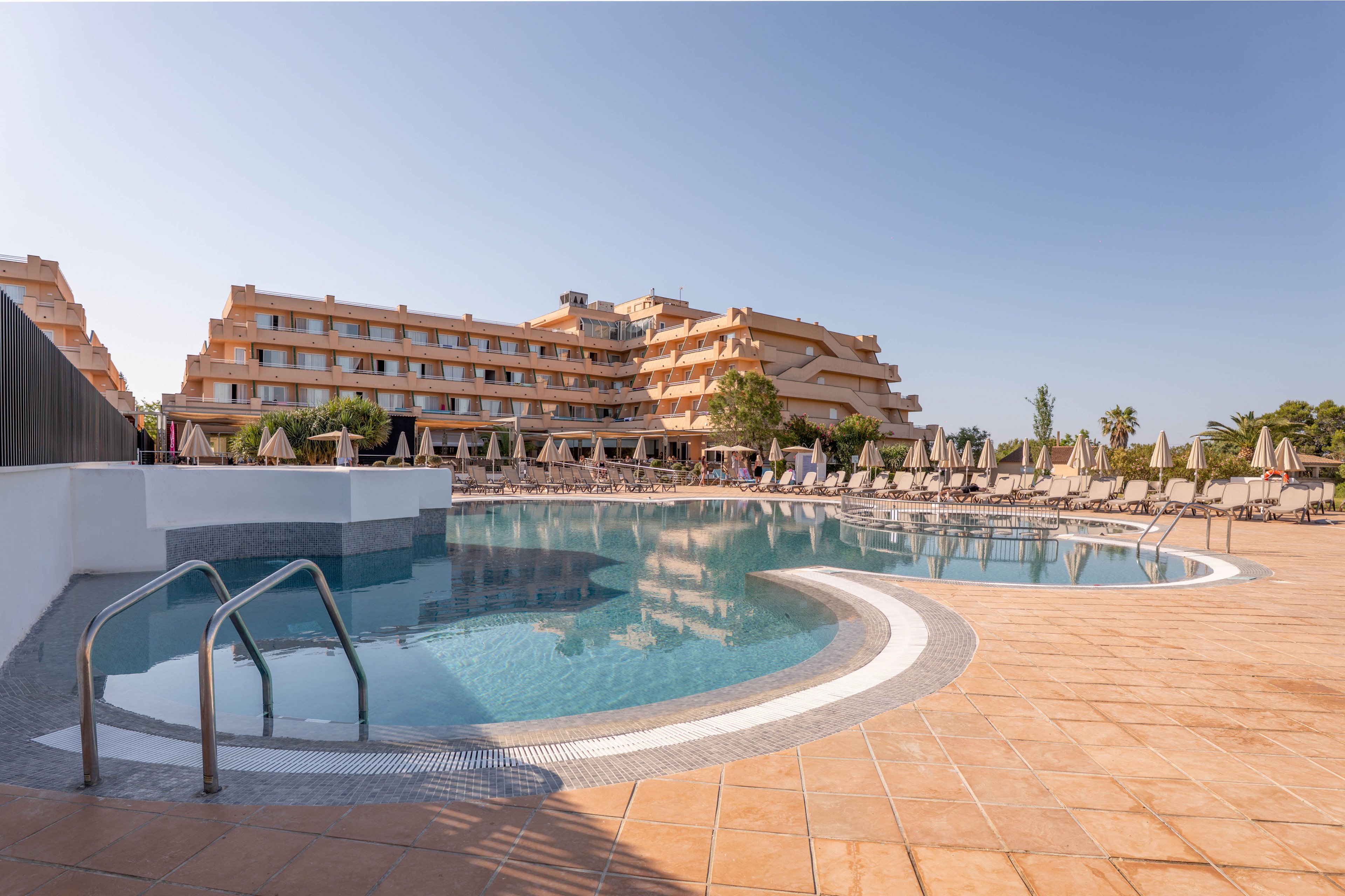 A large resort hotel overlooks a curved outdoor swimming pool on a clear day. The poolside area is lined with numerous sun loungers and umbrellas arranged in neat rows. Warm-toned tiles surround the water, reflecting the soft beige and orange hues of the multi-story building. The scene suggests a calm vacation atmosphere with no people currently using the facilities.