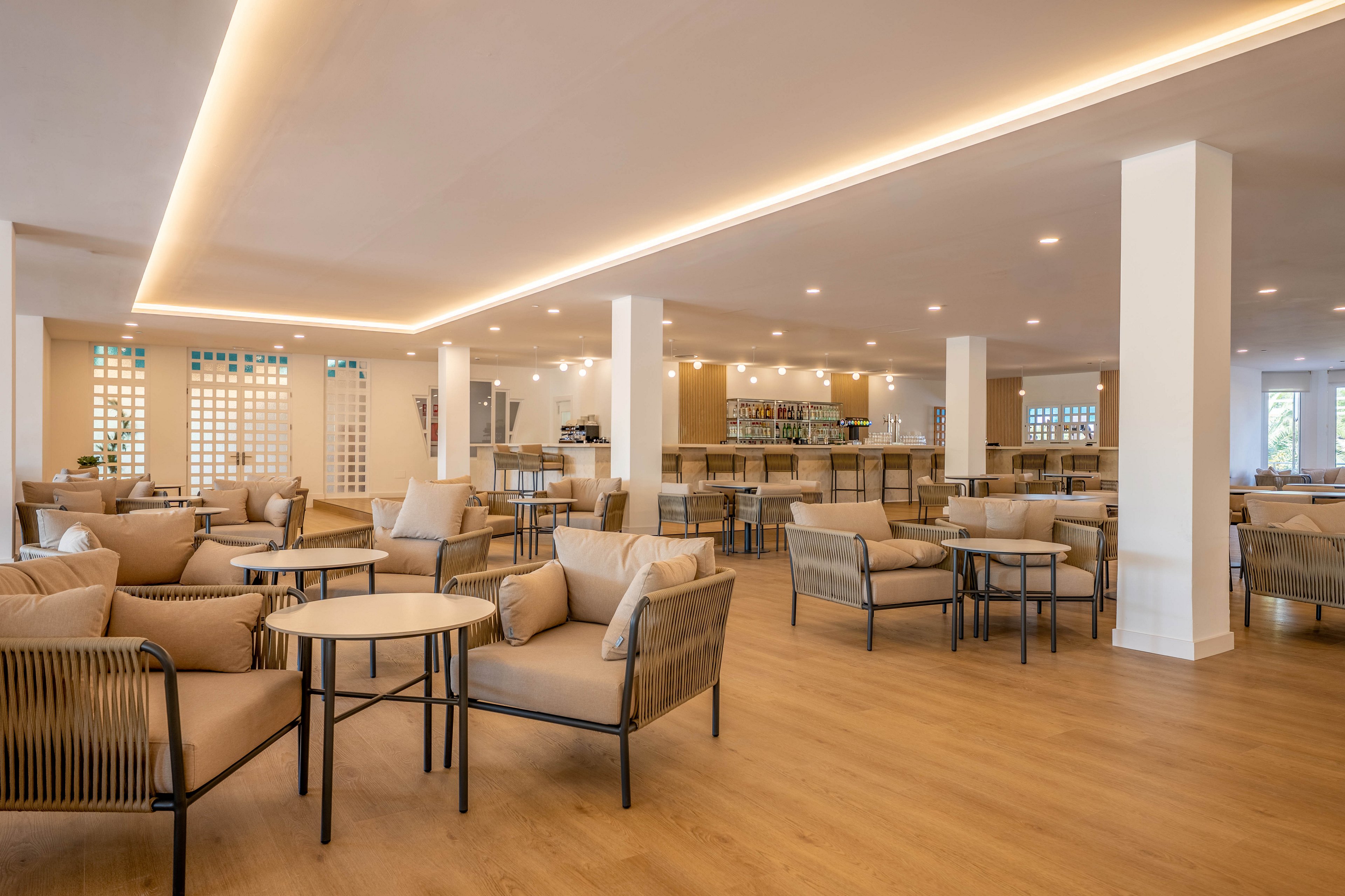Spacious contemporary lounge area features multiple seating groups with cushioned armchairs and round tables. The setting appears to be a hotel or resort bar with a long counter and shelves in the background. Warm wood flooring and recessed ceiling lights create a cozy, inviting atmosphere. Neutral beige and cream tones dominate the interior design, emphasizing comfort and relaxation.