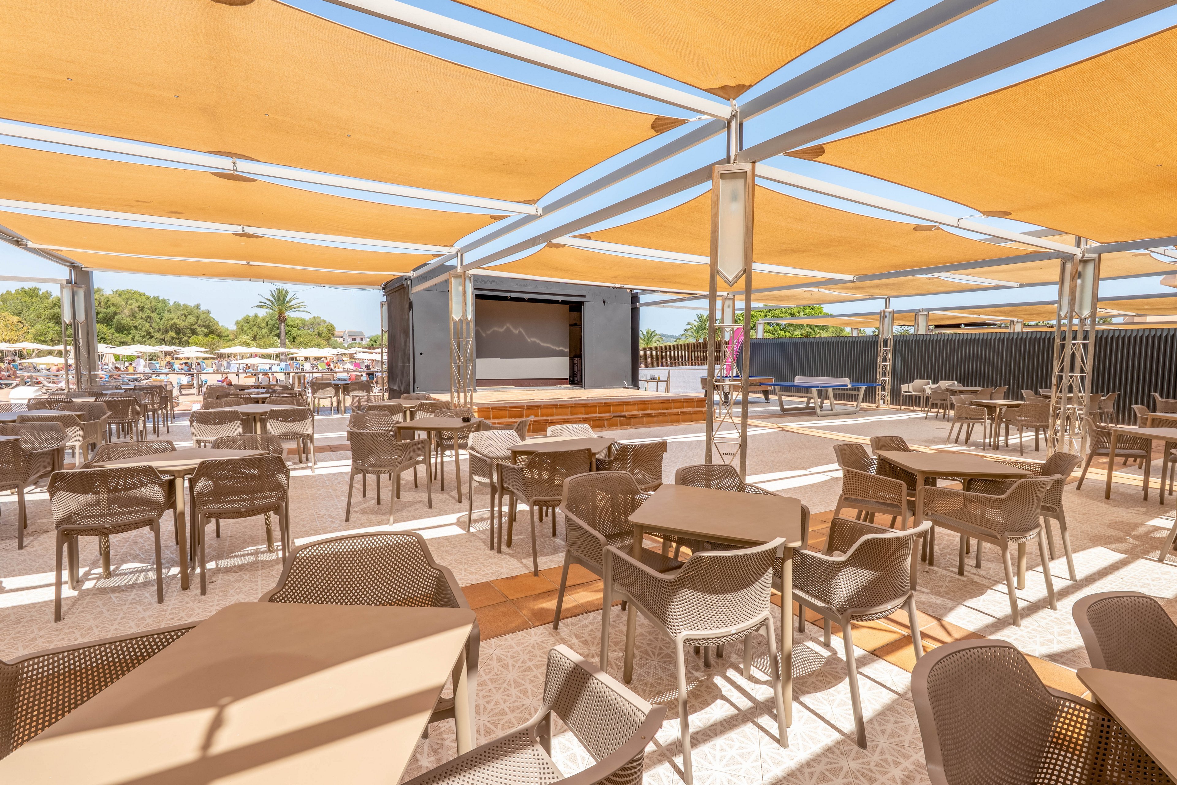 An outdoor cafe terrace features rows of empty tables and chairs under large beige shade canopies. In the background, a stage or large screen structure is set up for entertainment or presentations. Bright sunlight and clear skies create a warm, summery atmosphere, with greenery and people visible in the distant background. The scene suggests a relaxed resort or beachside dining and gathering area.