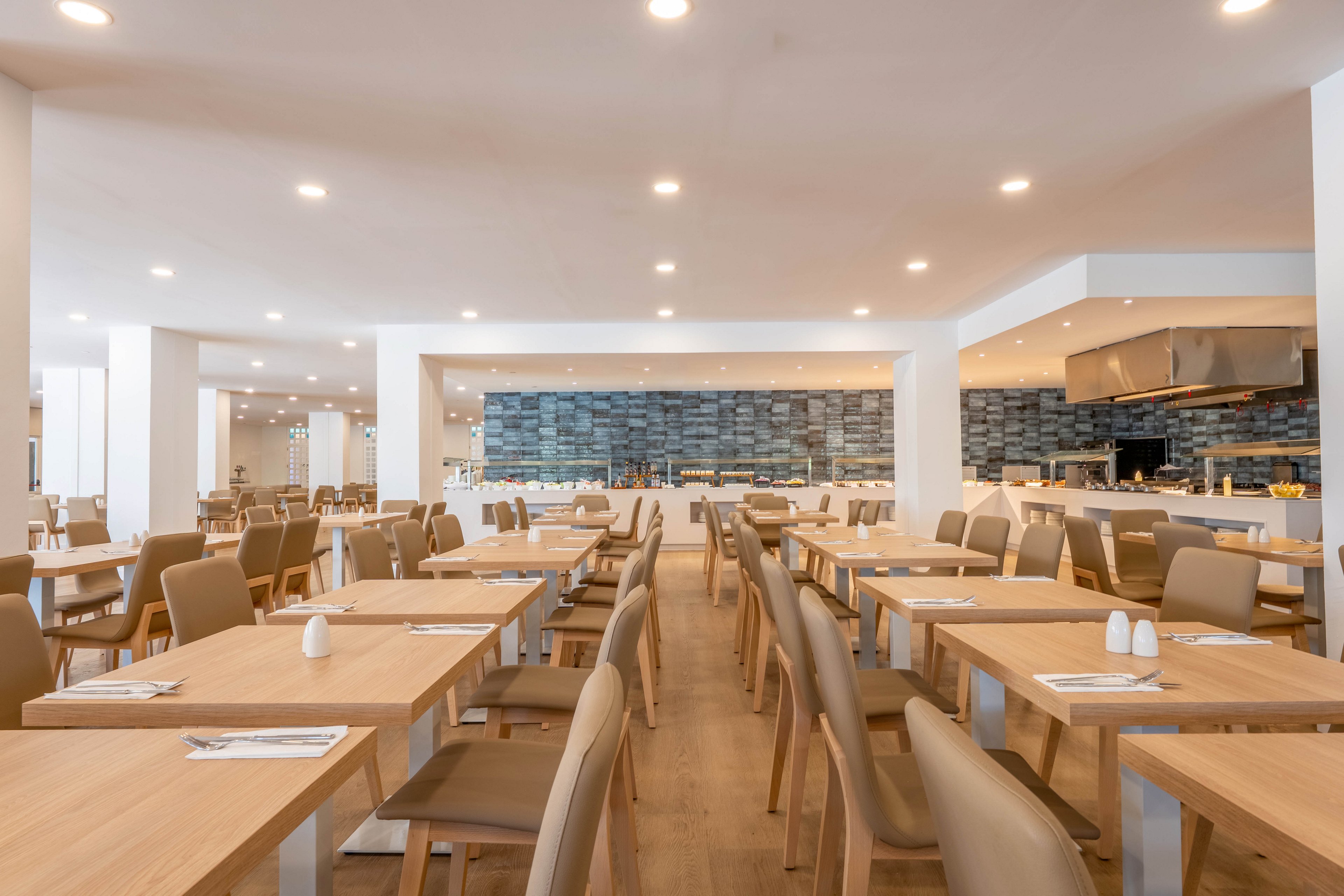 Spacious contemporary restaurant dining room with rows of light wooden tables and beige chairs. The room is brightly lit by numerous recessed ceiling lights, creating a clean and inviting atmosphere. Place settings with napkins and cutlery are arranged neatly on the tables, along with small salt and pepper shakers. In the background, a tiled wall and open kitchen or buffet area are visible, emphasizing a modern hospitality setting.