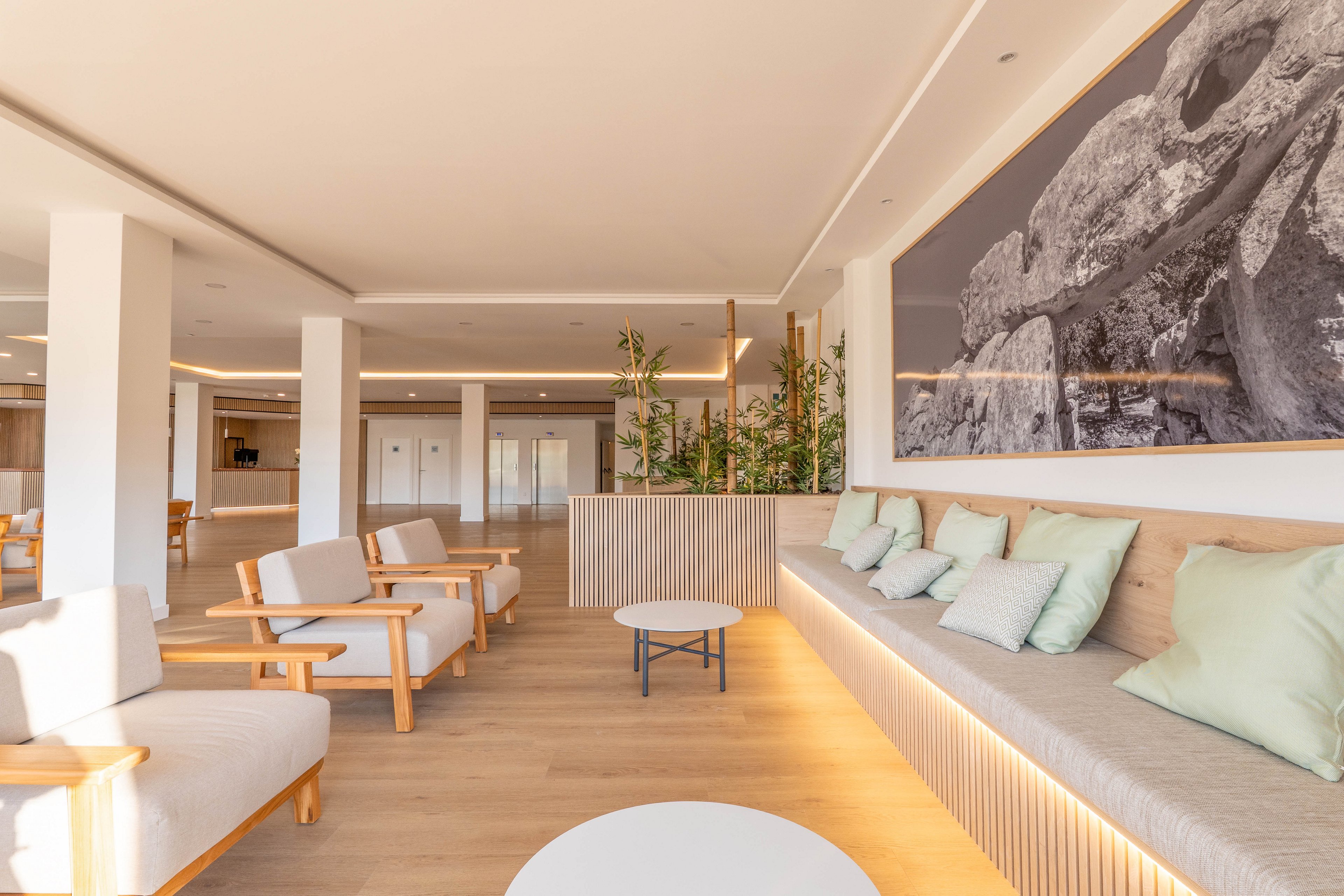 Spacious modern lounge area featuring light wood floors and neutral upholstered seating. The room includes armchairs, a built-in bench with pastel cushions, and small round coffee tables. Large columns and recessed ceiling lighting create a clean, open layout. A wide black-and-white landscape artwork and indoor plants add visual interest and a calming atmosphere.