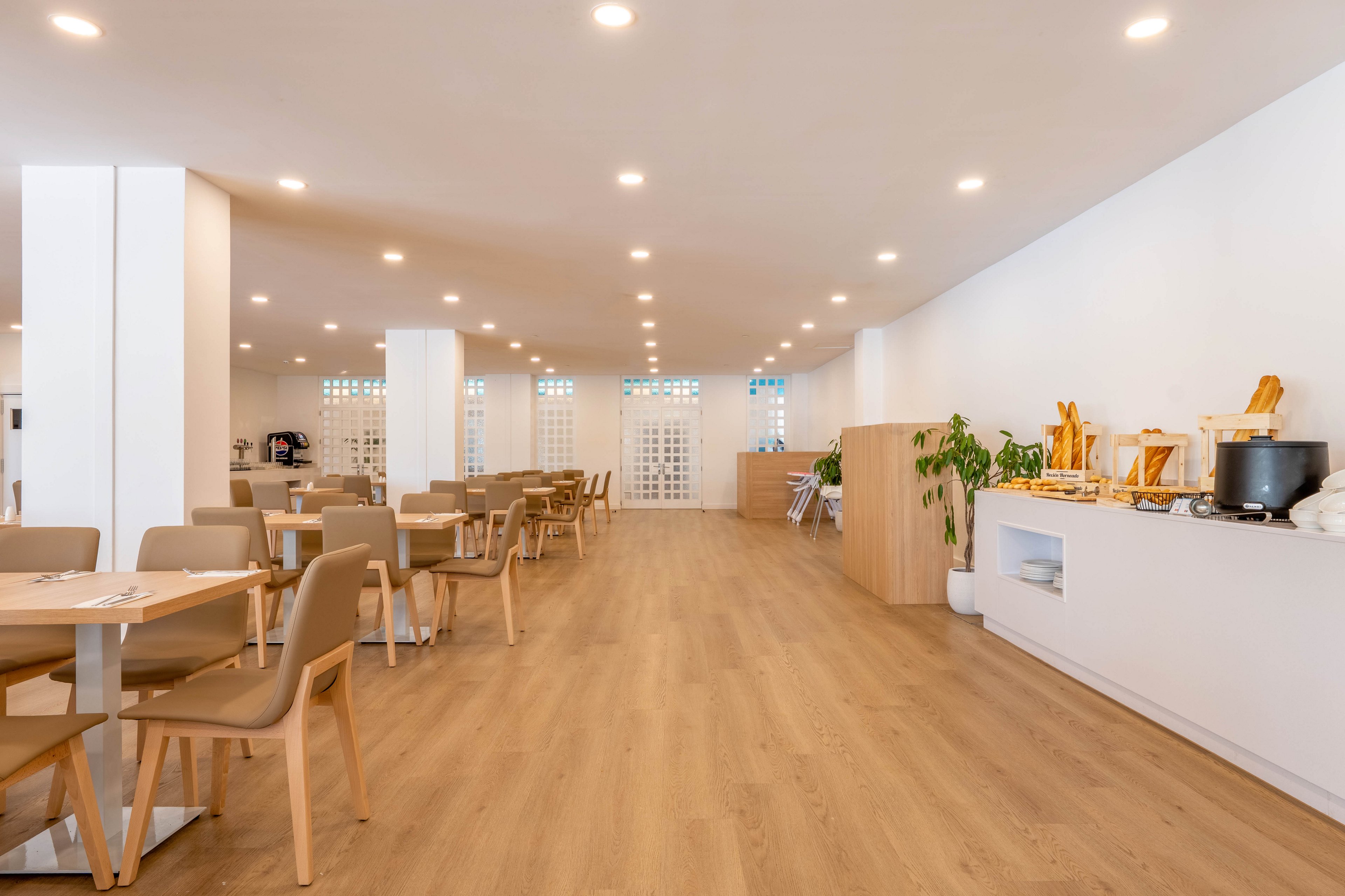 Spacious modern cafeteria interior with light wooden flooring and neutral-toned furniture. Several empty tables and chairs are arranged neatly throughout the room, creating an open and orderly dining area. On the right side, a breakfast buffet counter displays bread, pastries, and beverage dispensers alongside a potted plant. Recessed ceiling lights evenly illuminate the clean, minimalist space.