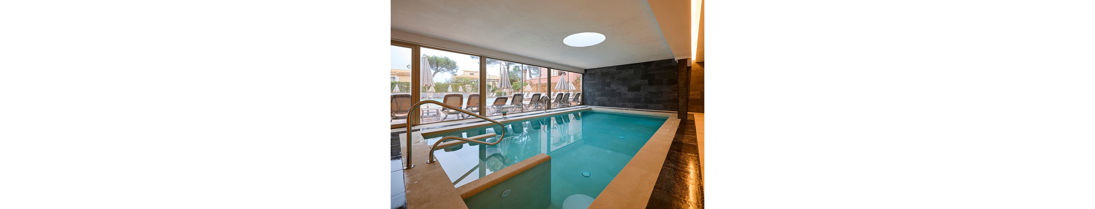Indoor swimming pool in a contemporary hotel or spa setting. Large floor-to-ceiling windows reveal an outdoor terrace with sun loungers and umbrellas. The pool features stainless steel handrails, clean beige tiling, and a dark stone accent wall. Soft natural light and warm interior tones create a calm, relaxing atmosphere.