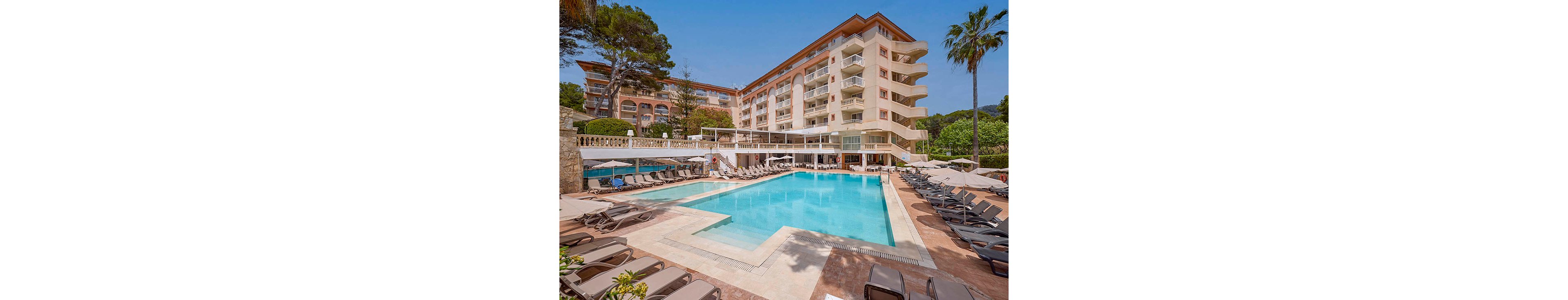 A large outdoor swimming pool sits at the center of a resort hotel complex. Multiple stories of balconies overlook the pool area, with sun loungers and umbrellas neatly arranged around the water. The scene is bright and sunny, suggesting a warm vacation destination surrounded by greenery and palm trees. The overall atmosphere is relaxed and inviting, ideal for leisure and holiday travel.