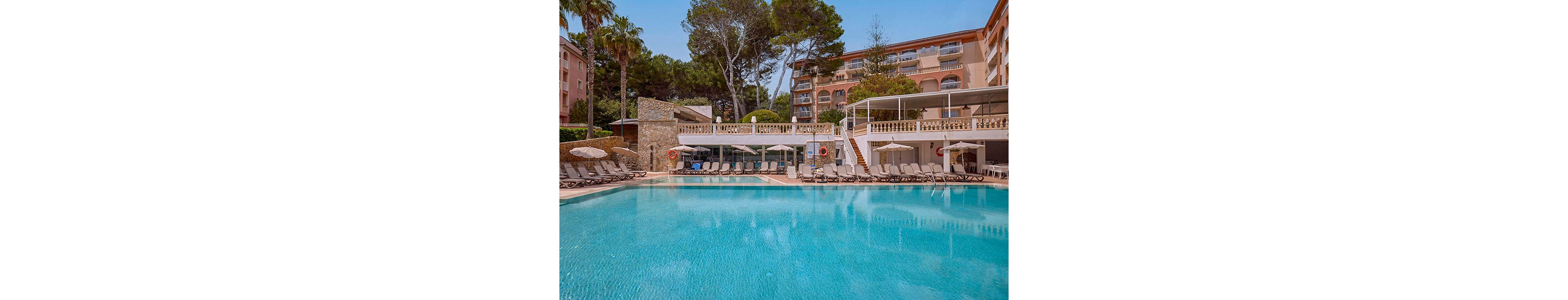 A large, clear blue swimming pool dominates the foreground of a sunny resort hotel. Rows of lounge chairs and umbrellas line the poolside terrace, suggesting a relaxing vacation setting. The multi-story hotel building with balconies and surrounding tall trees create a secluded, tranquil atmosphere. Warm tones of the architecture contrast with the cool turquoise water under a clear sky.