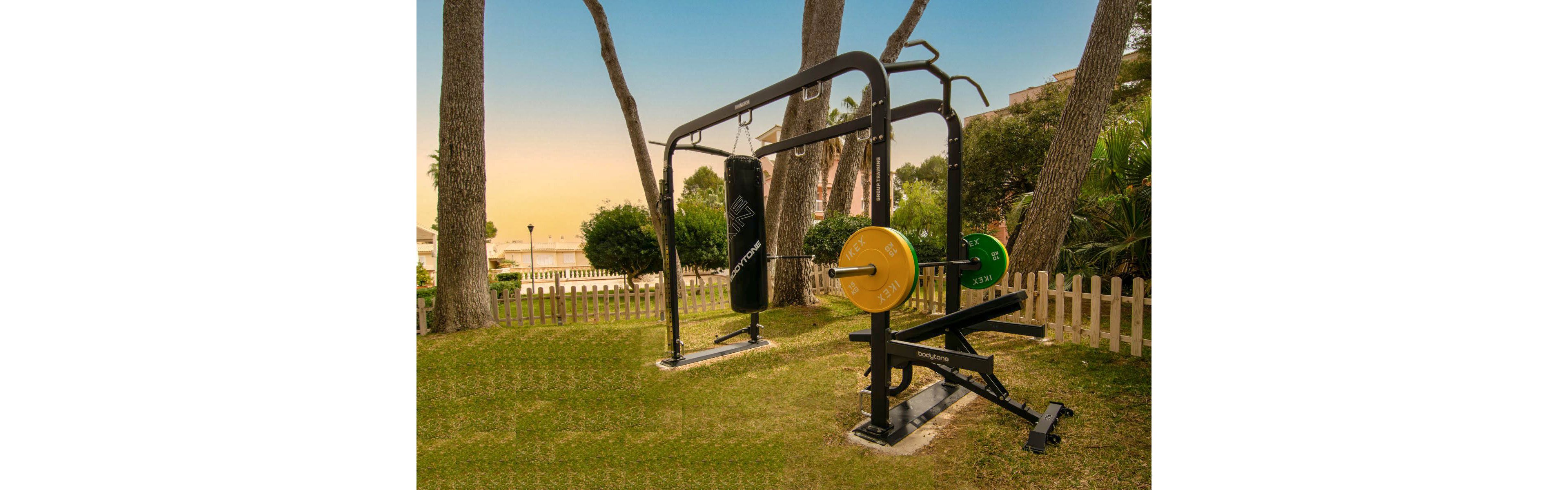 An outdoor gym station stands on a grassy lawn surrounded by trees and a white picket fence. The equipment includes a punching bag and a weightlifting bench with colorful weight plates. In the background, buildings and a distant waterfront are visible under a warm evening sky. The scene combines fitness equipment with a tranquil, resort-like environment.