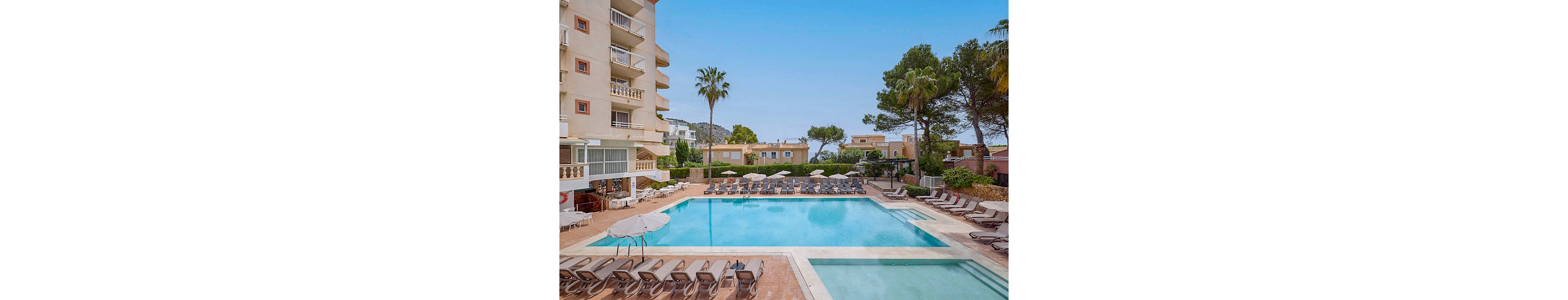 A large outdoor swimming pool is surrounded by lounge chairs and umbrellas in the courtyard of a multi-story hotel. The clear blue water reflects the bright daytime sky, creating a relaxing vacation atmosphere. Tall palm trees and other greenery frame the pool area, while additional residential-style buildings are visible in the background. The scene is calm and inviting, with no people present.