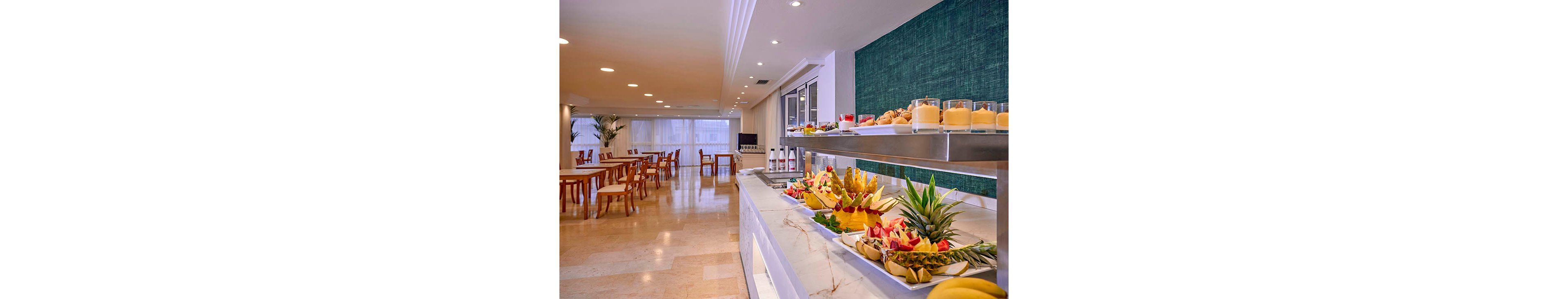 A hotel dining room features a well-organized breakfast buffet with an emphasis on fresh fruit. Sliced pineapple, watermelon, papaya, and other fruits are neatly arranged on white platters along a marble counter. Glasses and bowls of juices or smoothies are lined up on the upper shelf beside dispensers and condiments. In the background, wooden tables and chairs sit in a bright, spacious room with large windows and recessed ceiling lights.
