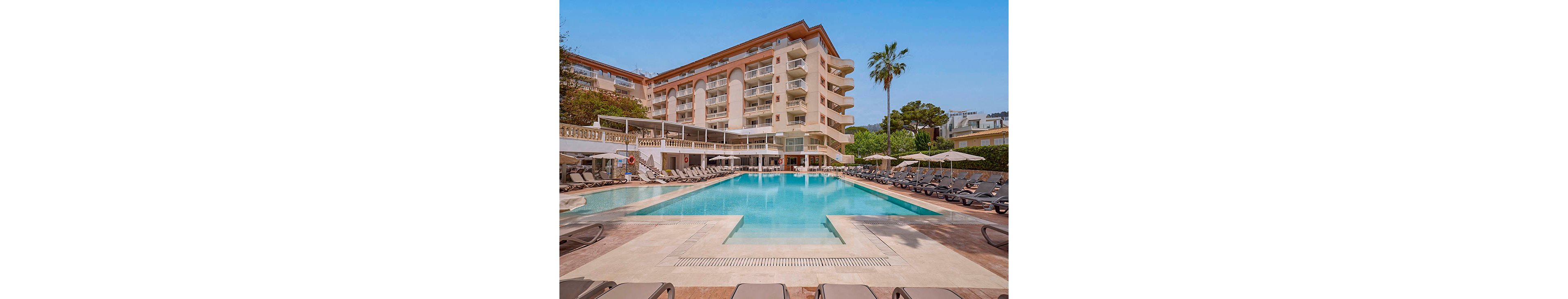 A large outdoor swimming pool sits at the center of a luxury hotel courtyard. Multiple stories of balconies overlook the pool, each with railings and shaded areas. Numerous sun loungers and umbrellas line the poolside, suggesting a relaxing vacation setting. A tall palm tree and surrounding greenery add to the resort atmosphere under a clear blue sky.