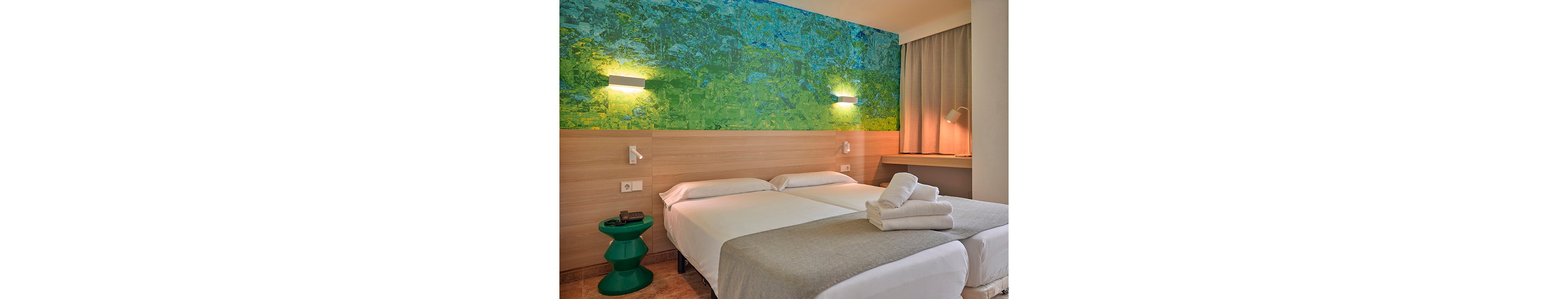A contemporary hotel room features two neatly made twin beds with light gray blankets and white linens. Freshly folded white towels are placed on one of the beds, suggesting readiness for new guests. The room is accented by a vibrant green and blue wall mural above a light wood headboard and a unique green side table. Warm wall and desk lamps create a cozy, inviting atmosphere next to a curtained window and simple work surface.