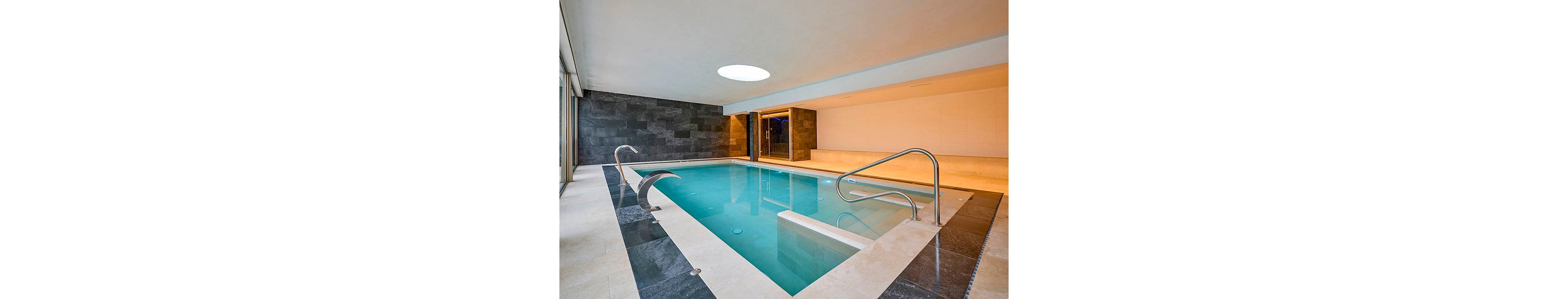 A sleek indoor swimming pool is featured in a contemporary spa or residential setting. The pool is surrounded by light stone tiles and dark slate wall panels, creating a clean, minimalist look. Stainless steel handrails and a curved water feature add a touch of luxury. A circular skylight and recessed ceiling lights illuminate the calm turquoise water, emphasizing the tranquil atmosphere.
