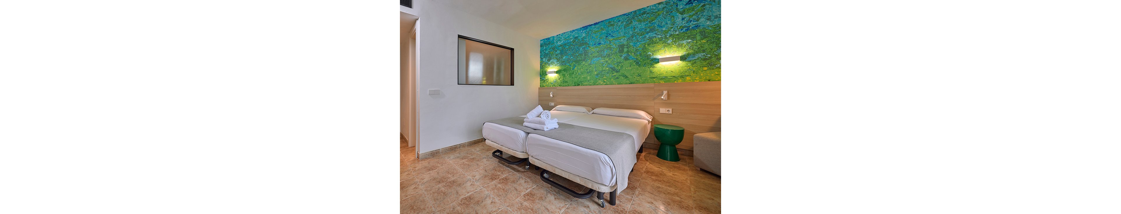 A contemporary hotel room features two neatly made twin beds pushed together with folded white towels placed on top. The space has light-colored tiled flooring and a wooden headboard that runs the length of the beds. A vibrant abstract wall mural in blue and green tones adds color above the headboard, complemented by minimalist wall-mounted lights. A small green side table and a partial view of a sofa complete the clean, modern interior.