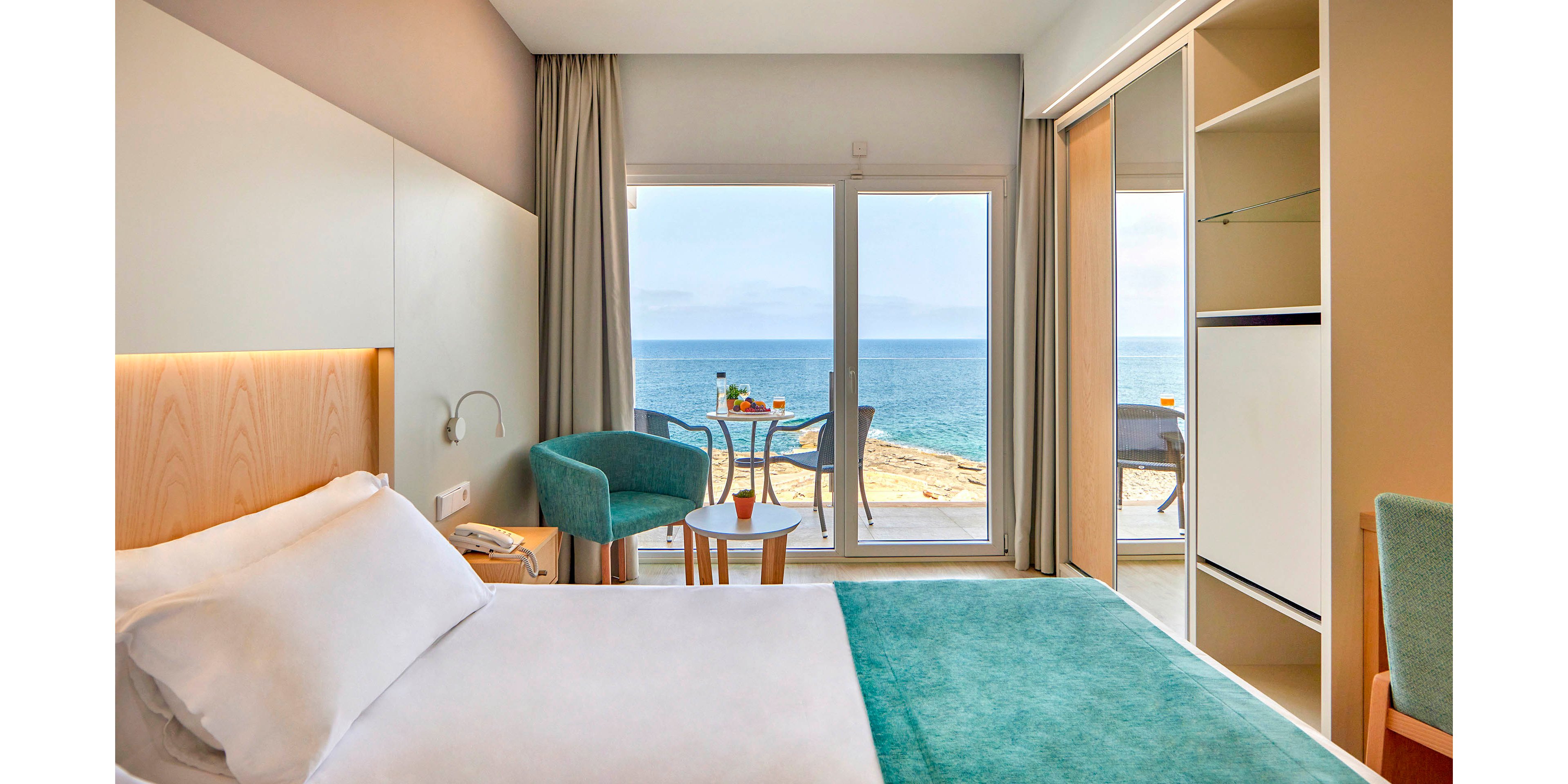 A modern hotel room features a neatly made bed with white linens and a teal blanket. Large glass doors open to a balcony with a small round table, two chairs, and a view of the sea and rocky shoreline. Soft neutral tones, teal upholstery, and warm accent lighting create a calm, inviting atmosphere. Built-in storage and minimalist decor emphasize a clean, contemporary style.