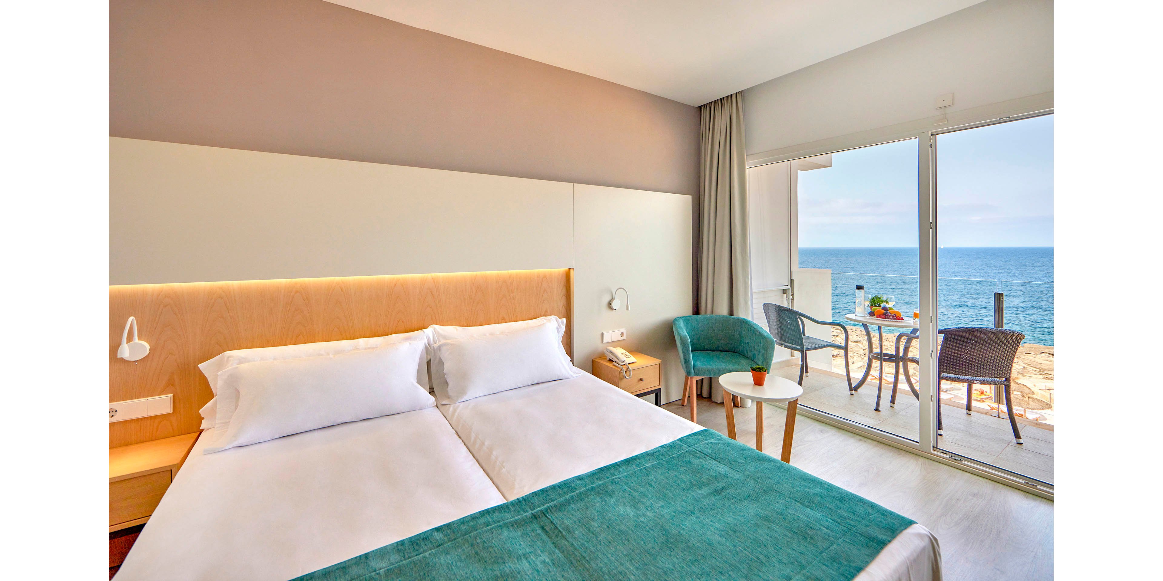 A bright, modern hotel room features two neatly made twin beds with white linens and a teal throw. The room opens onto a balcony with a clear view of the ocean and blue sky. Contemporary furniture, including a small round table and teal armchair, adds comfort near the sliding glass doors. Natural light fills the minimalist interior, creating a calm and inviting coastal atmosphere.