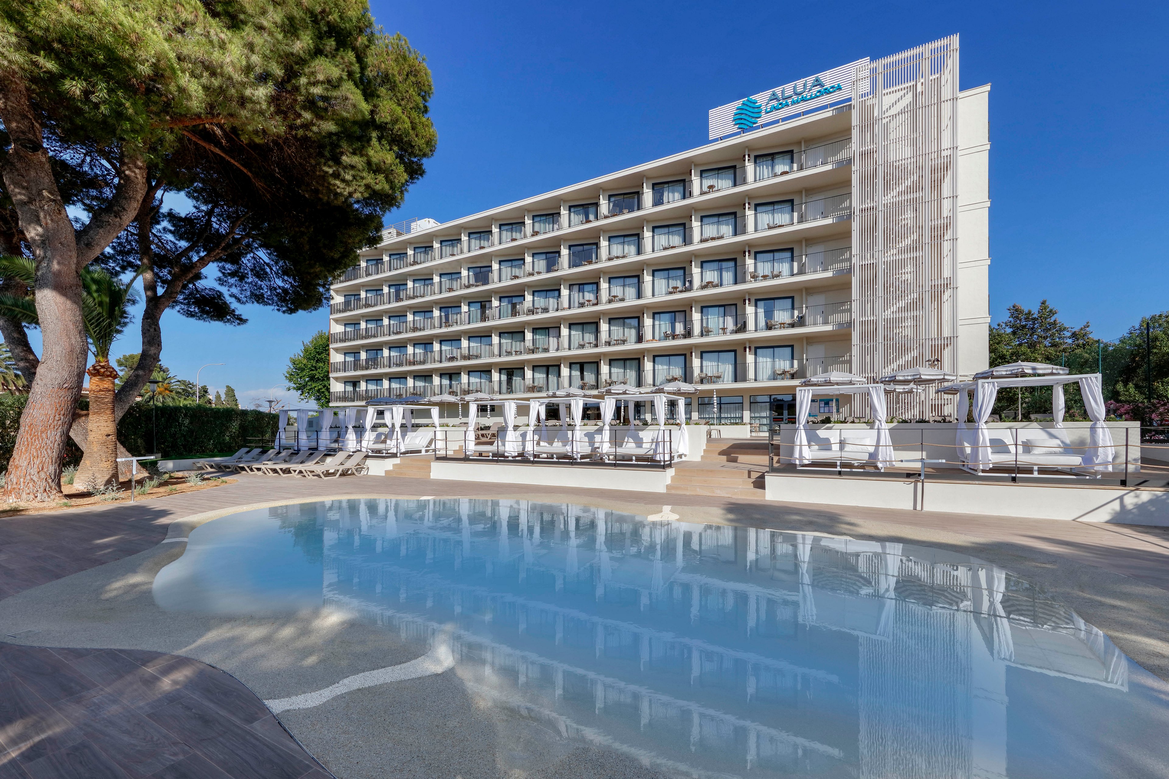 A contemporary multi-story hotel stands behind a calm outdoor swimming pool with clear blue water. White sun loungers and canopy daybeds line the poolside deck, creating a relaxed vacation atmosphere. Tall trees frame the scene, and the sky is bright and clear, suggesting a sunny day. The building facade features balconies on each floor and a visible hotel name at the top.